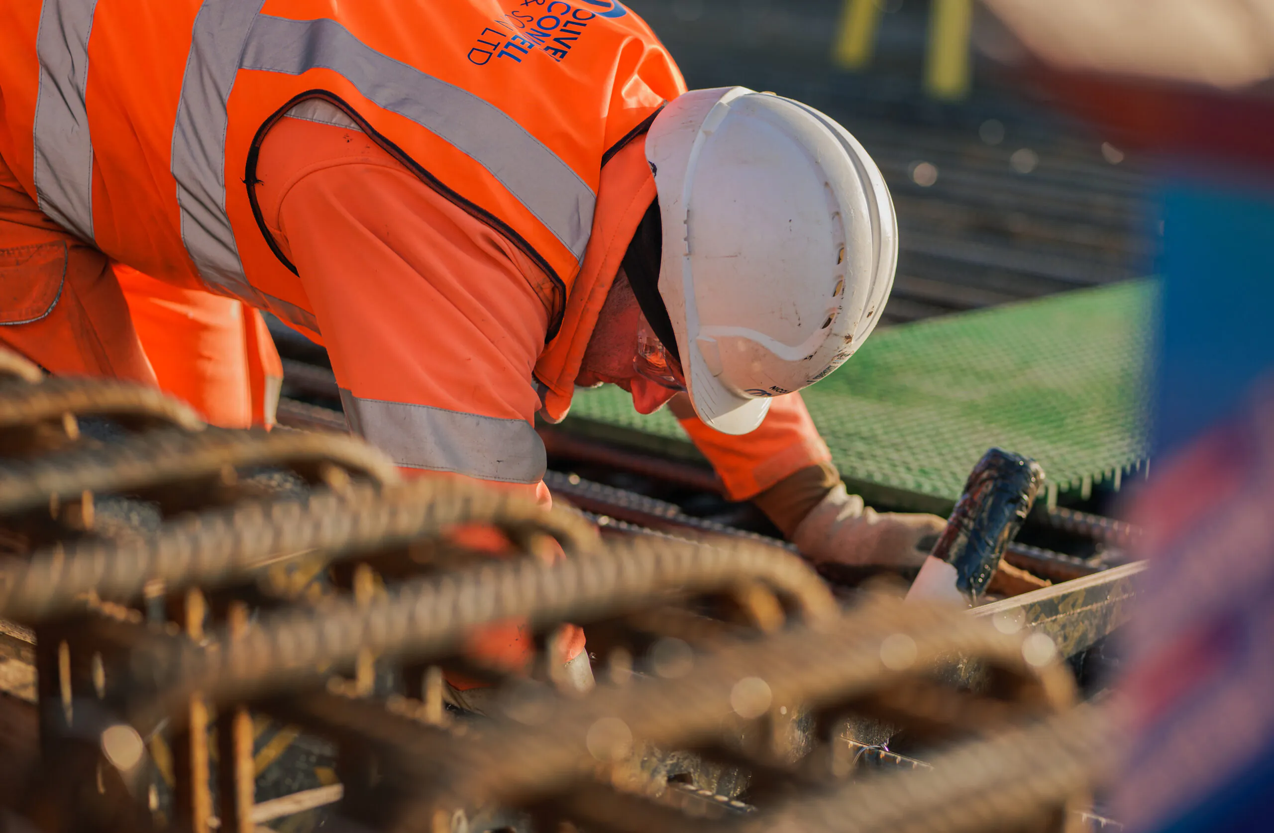 Worker in high-visibility clothing and hard hat bending over rebar at a construction site during the day.