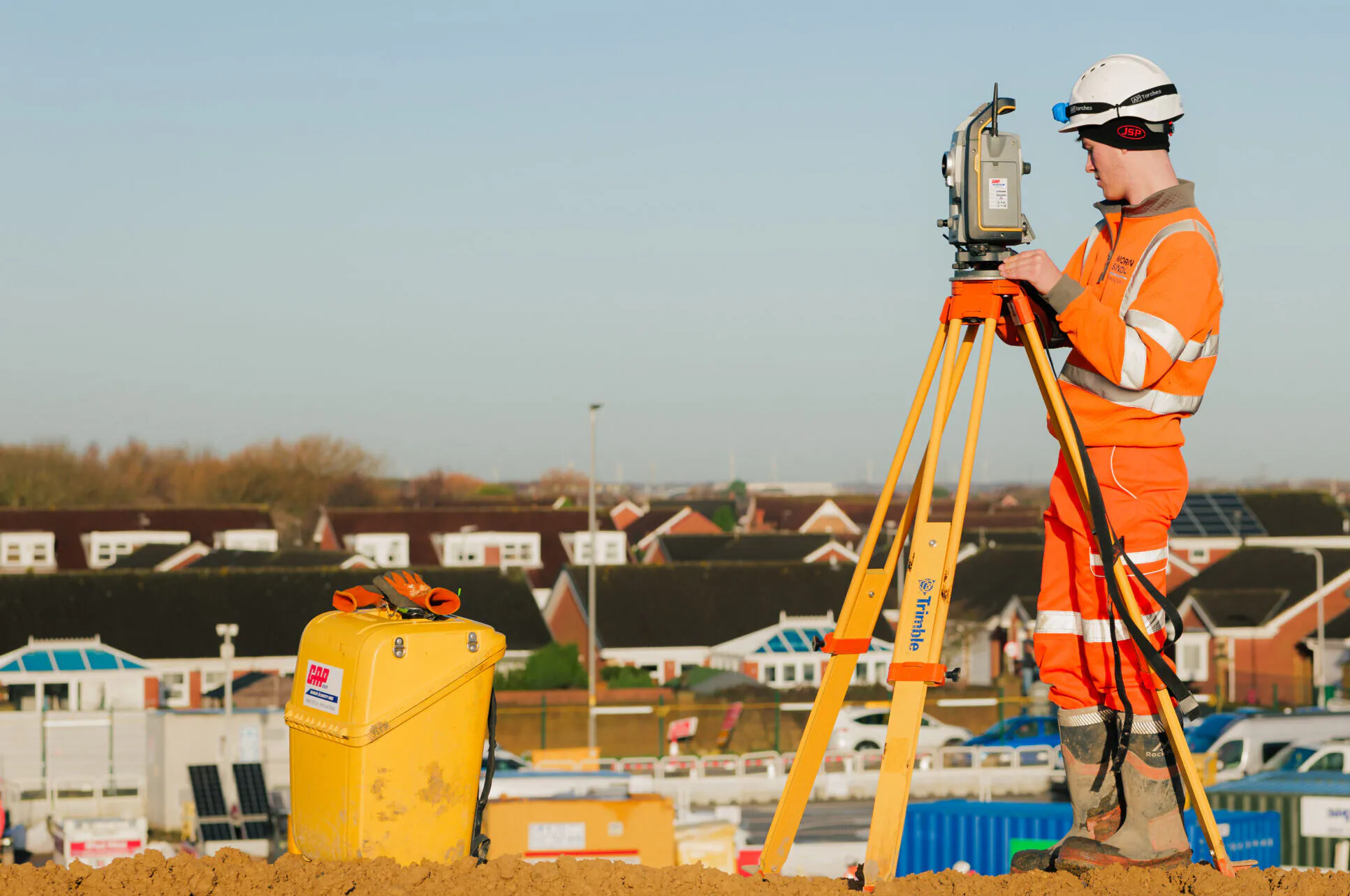 Surveyor in high-visibility clothing using tripod-mounted equipment at an outdoor construction site during daylight.
