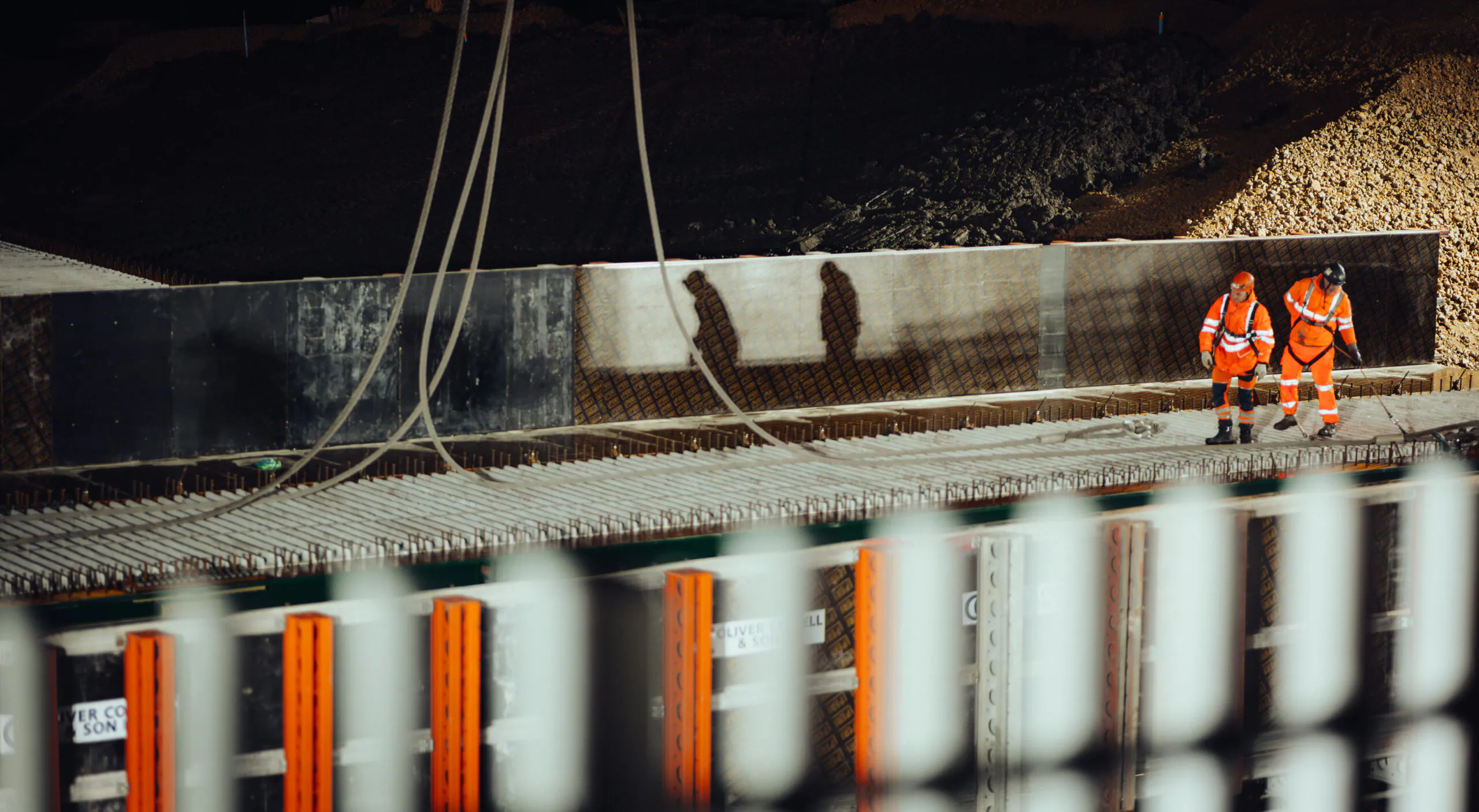 Two workers in high-visibility clothing walking along elevated rebar at night, casting shadows on the wall behind.