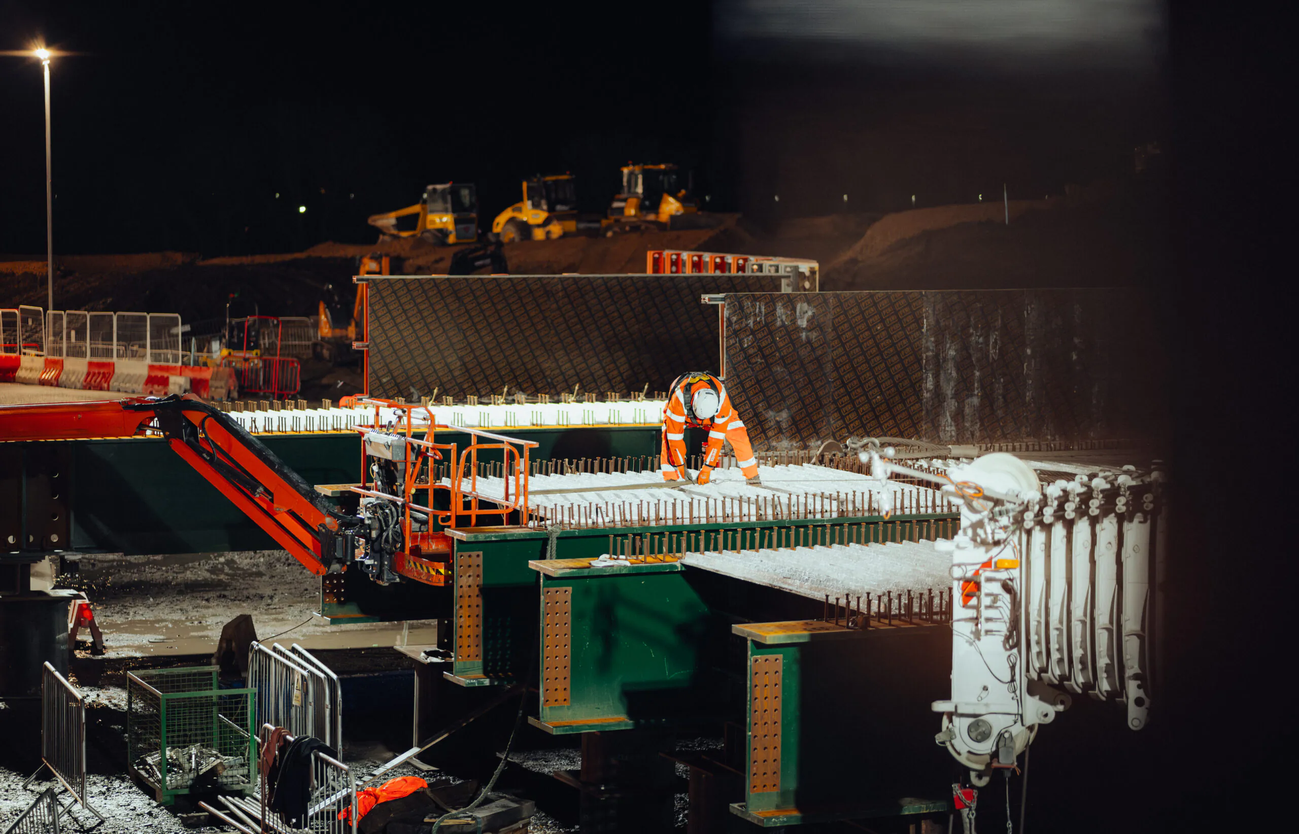 Nighttime construction work with workers in high-visibility clothing assembling steel structures under bright lights.