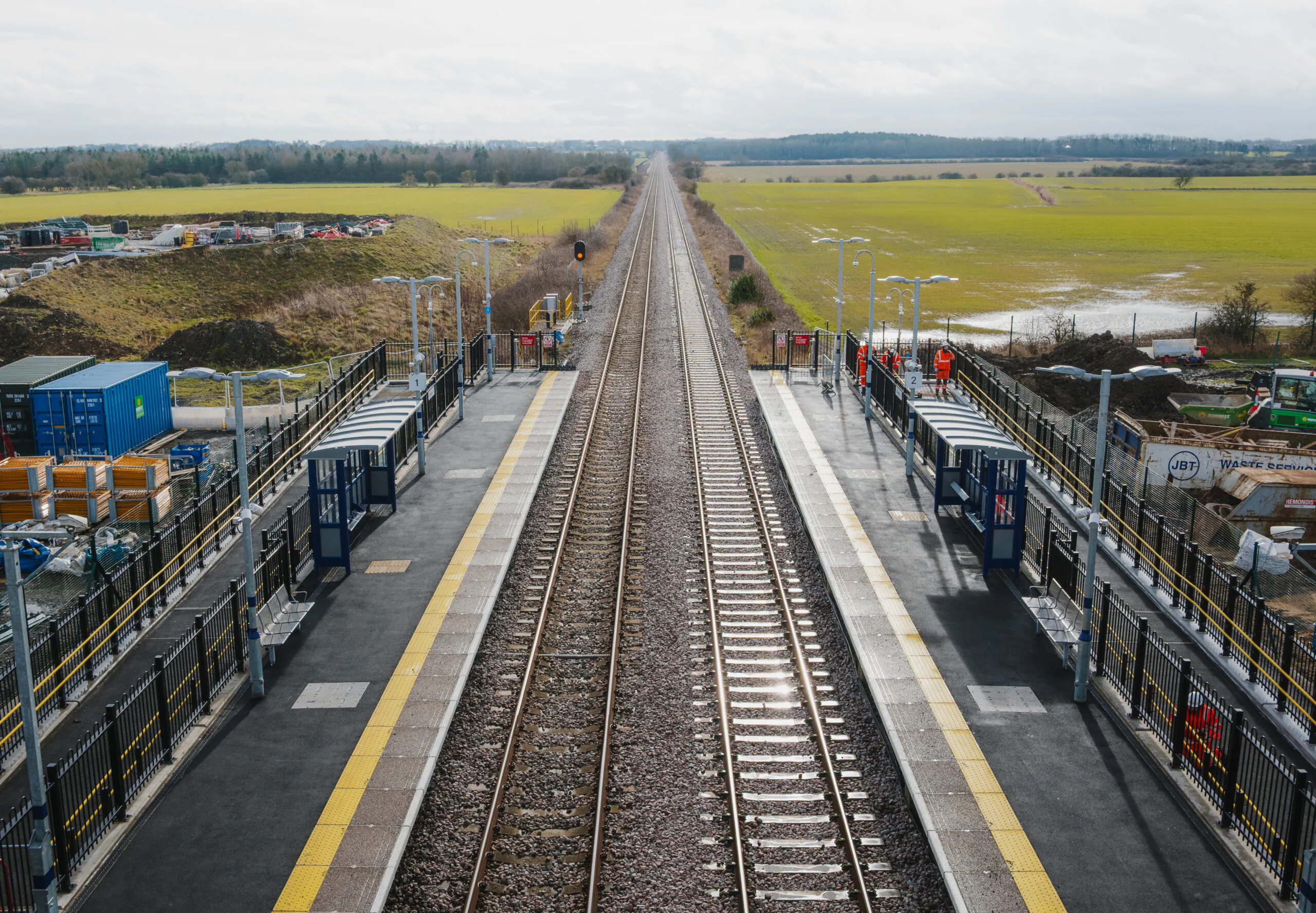 Aerial view of railway tracks flanked by platforms and fields under an overcast sky.