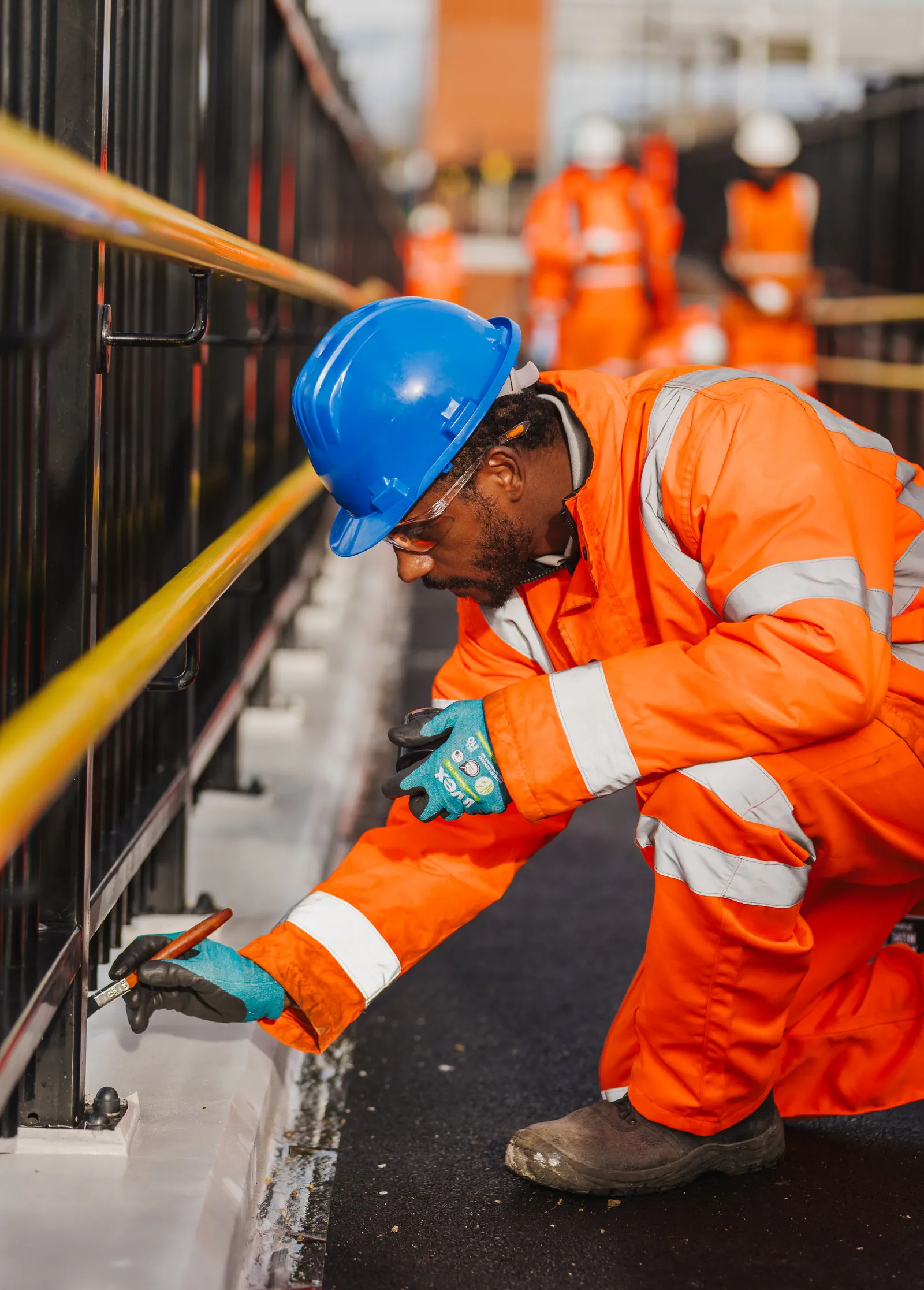 Worker in bright safety gear kneels beside a railing performing maintenance with hand tools.