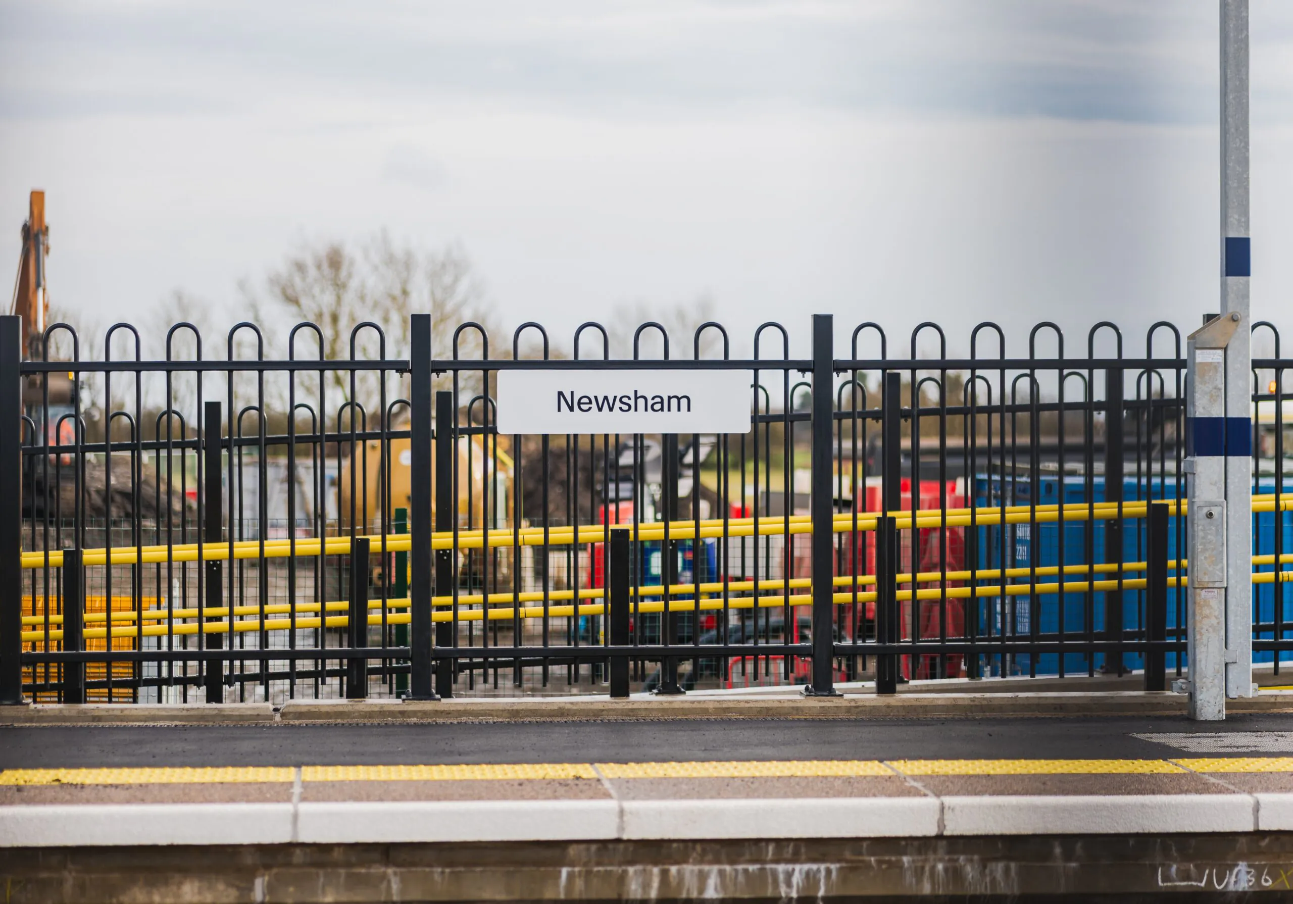 Close-up of the "Newsham" station sign mounted on black railings with construction equipment in the background.