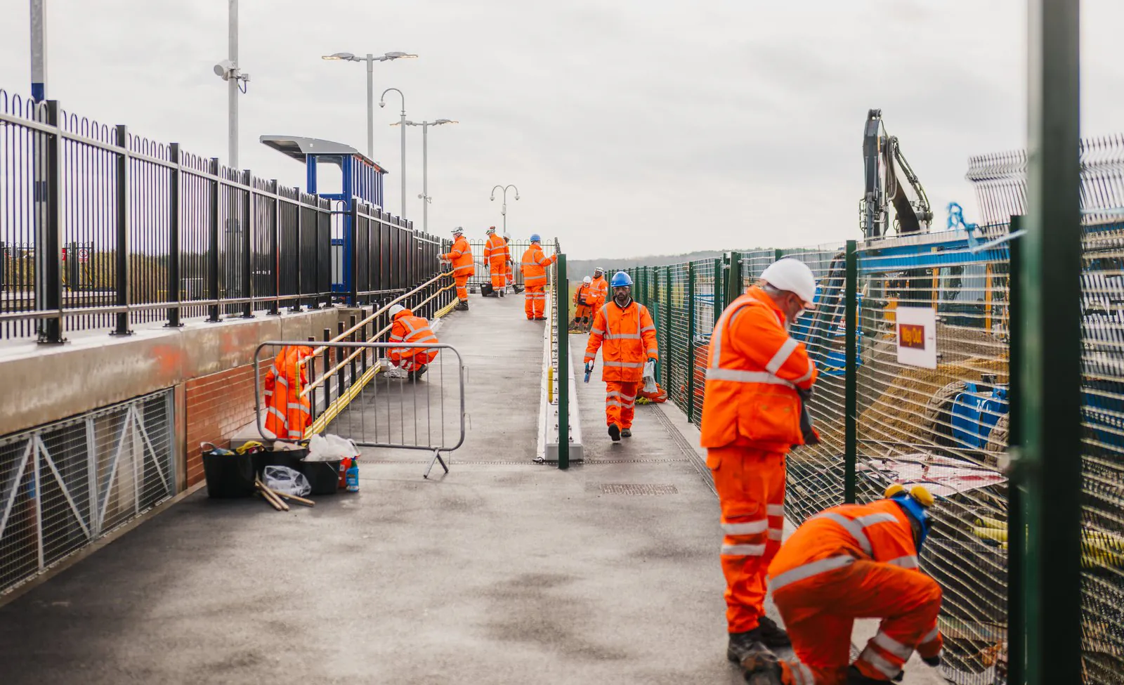 Workers in orange safety gear perform tasks along an elevated walkway at a construction or railway platform site.