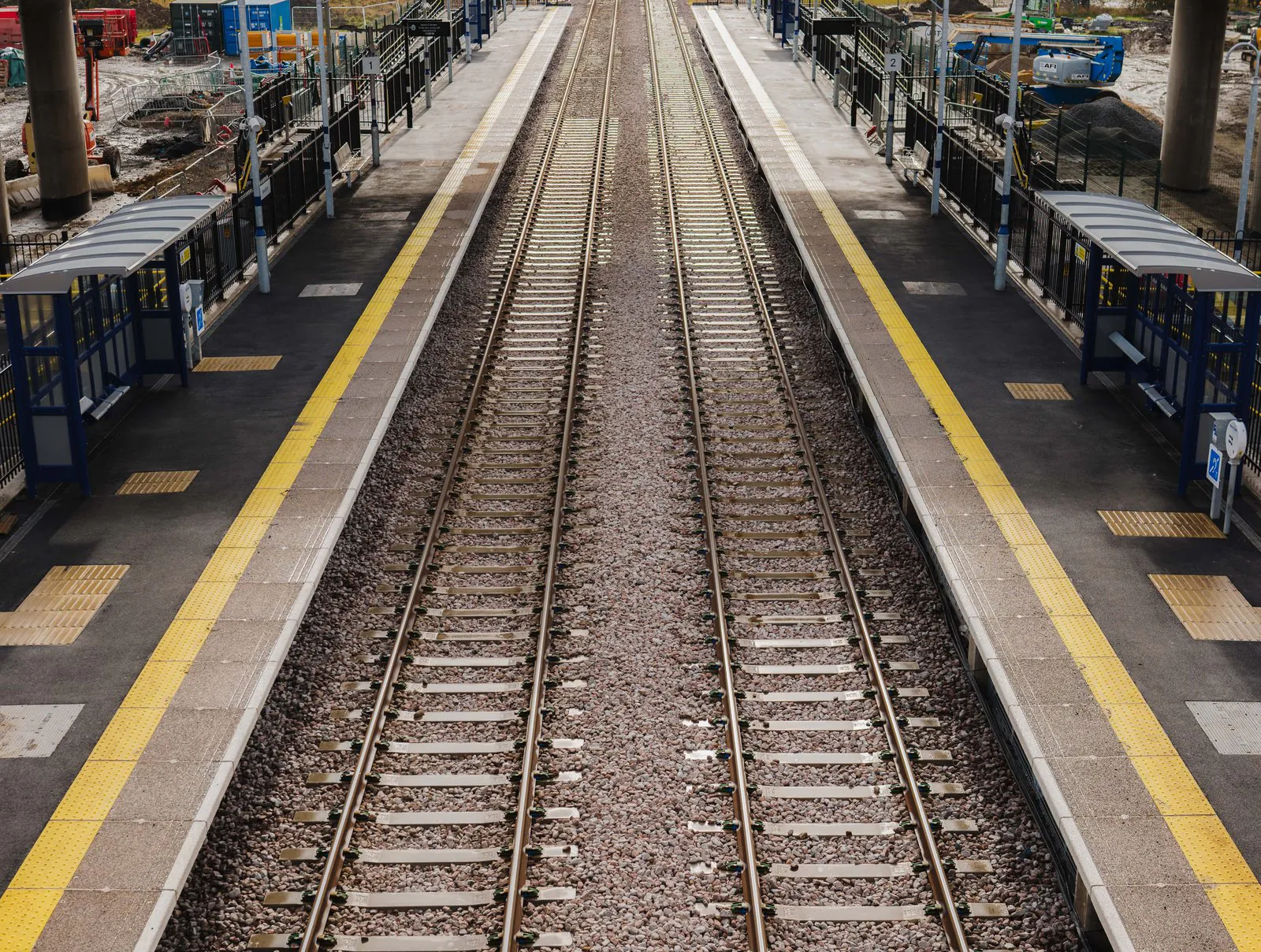 Empty railway station platform with dual tracks, yellow safety lines, shelters, benches, and signage visible along the platform.