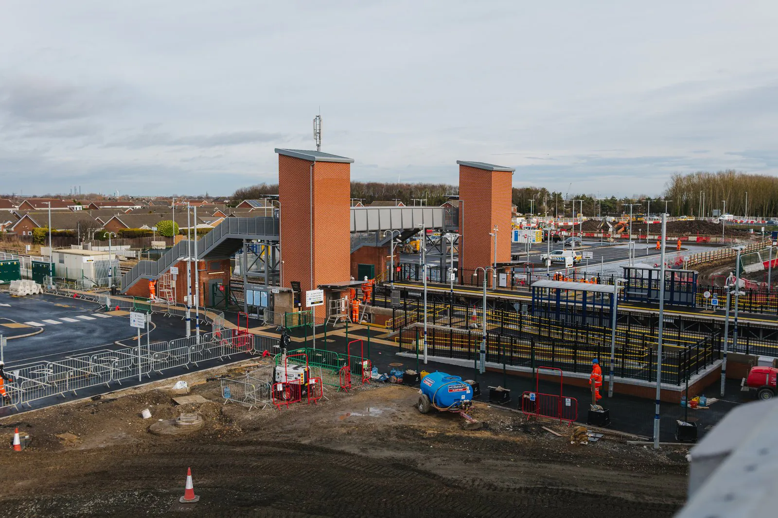 Construction site featuring two tall brick structures with scaffolding, workers in orange safety gear, and temporary fencing surrounding the area.