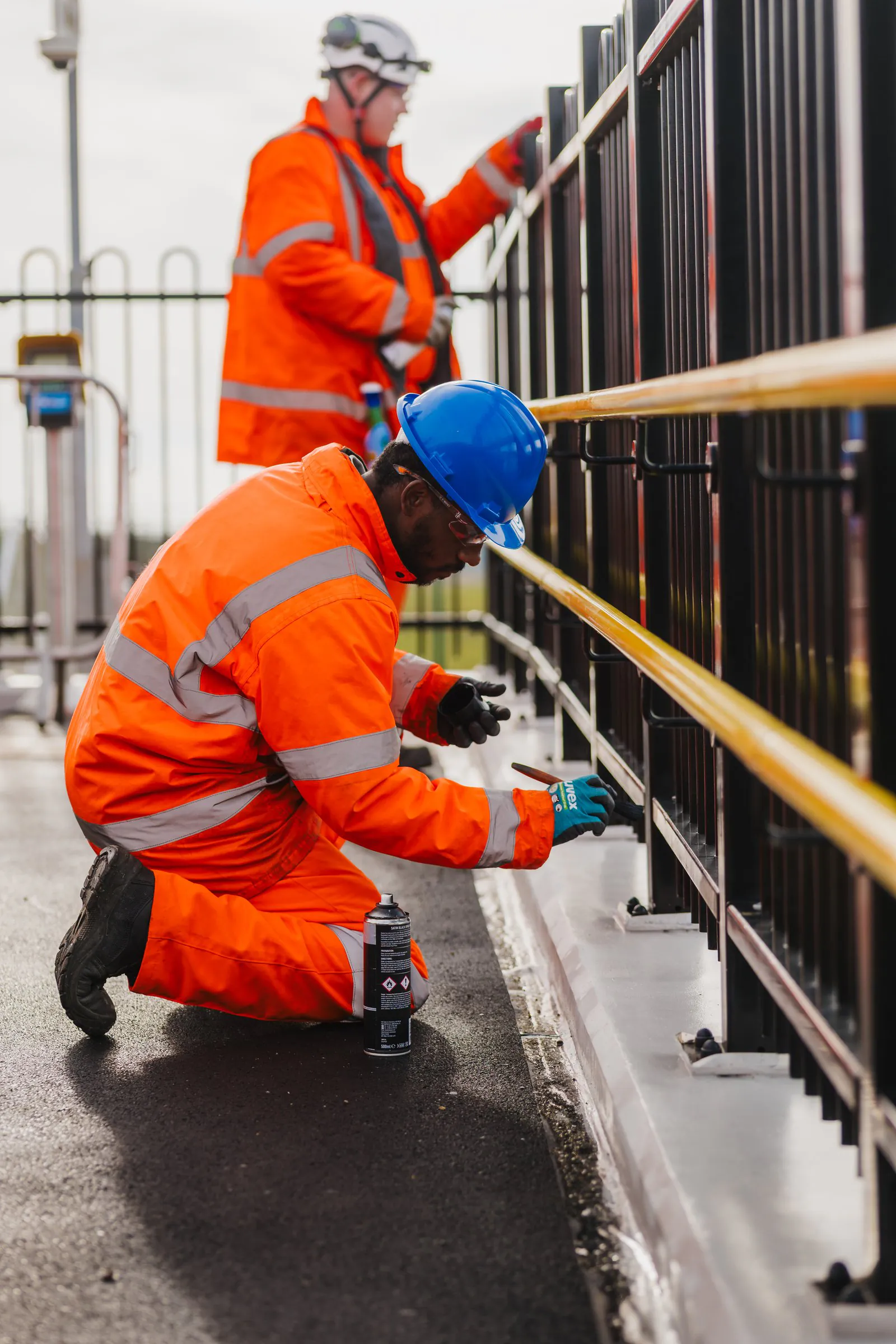 Worker in bright safety gear kneels beside a railing performing maintenance with hand tools.