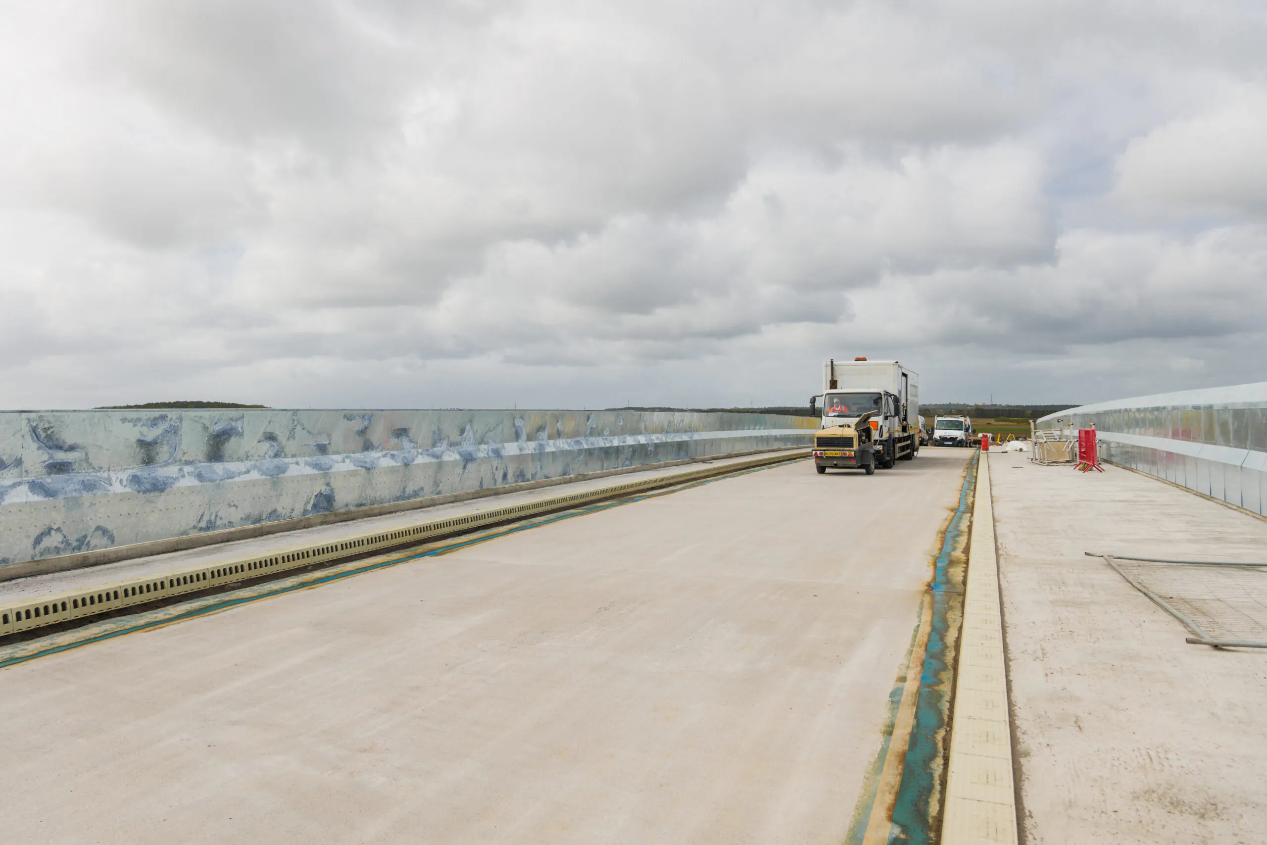 Construction site on an elevated roadway or bridge with two vehicles parked on it under an overcast sky.