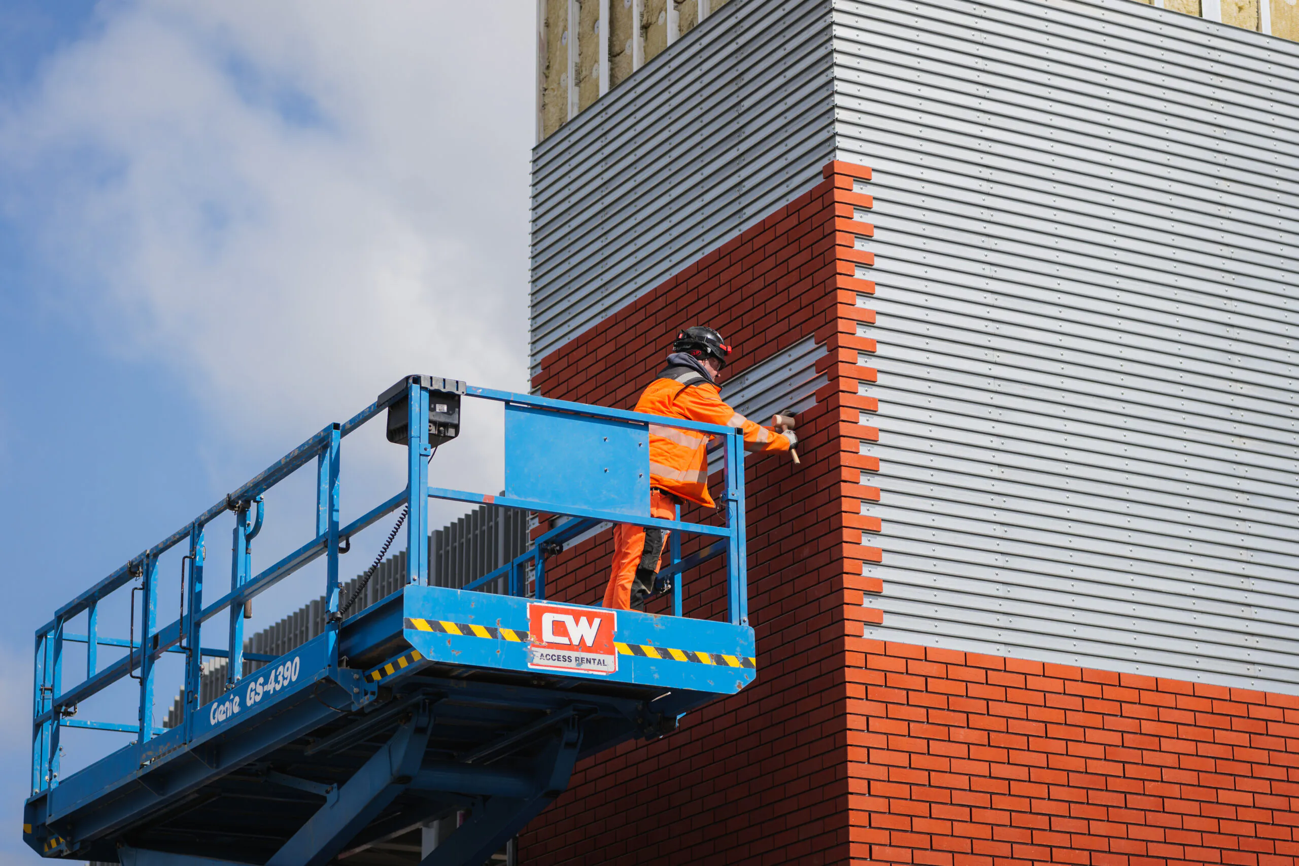 "Worker in high-visibility clothing installing or repairing exterior building panels from an elevated platform during daylight.