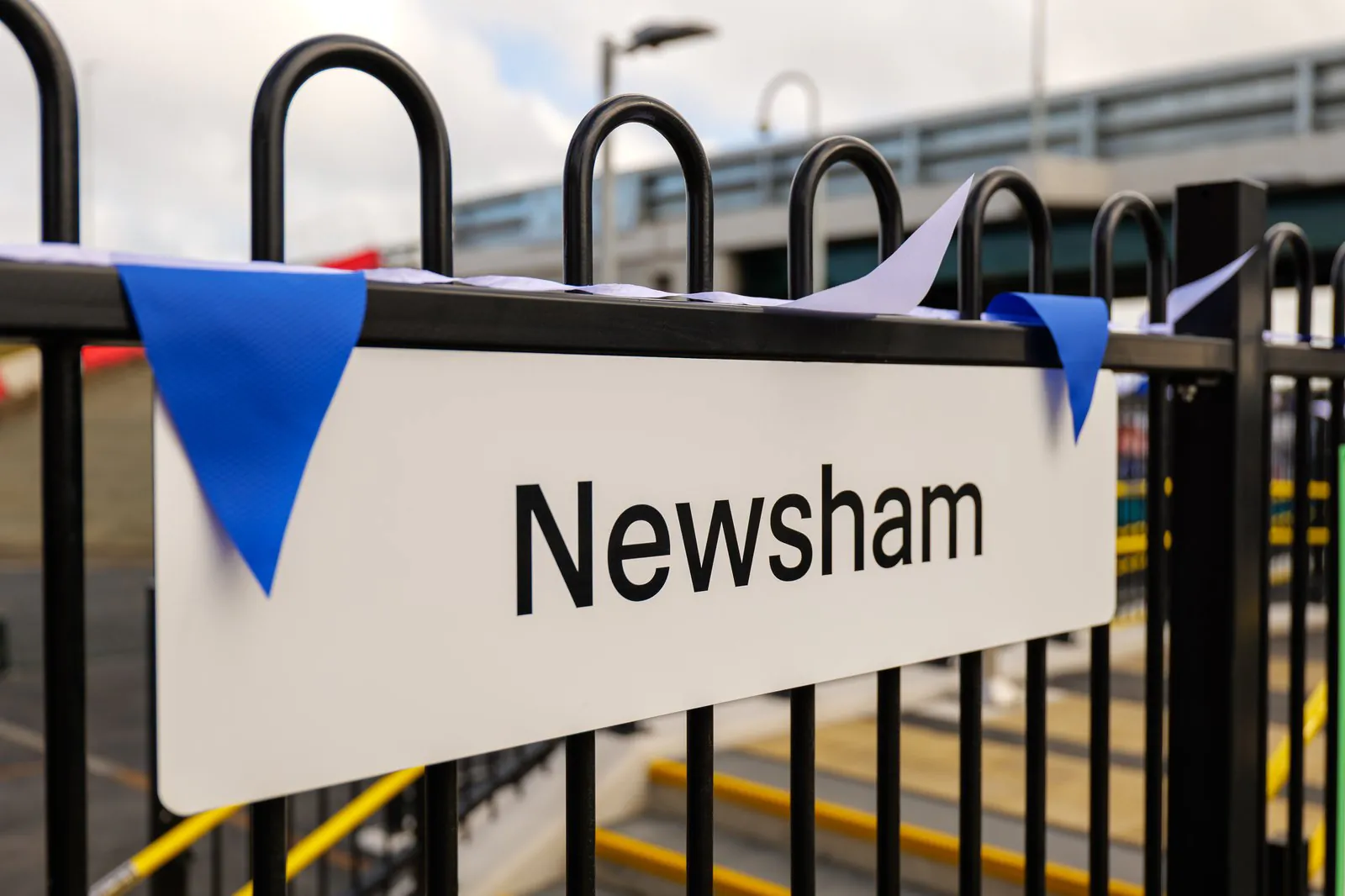 Close-up of a "Newsham" station sign mounted on a black metal fence, decorated with blue and white triangular bunting. Blurred elements of the railway station, including stairs and railings, are visible in the background.