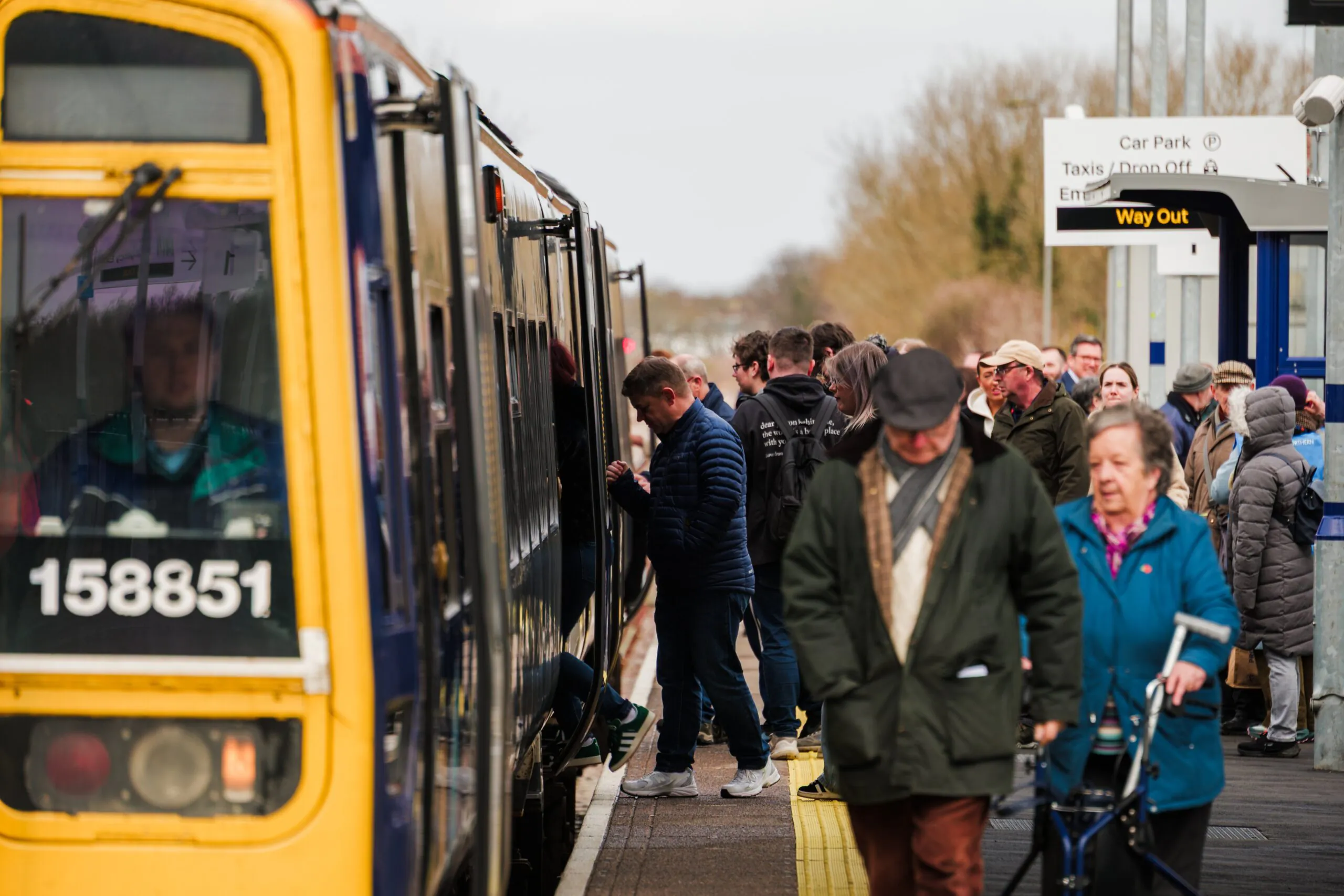Passengers board or exit a train marked "158851" at a station platform with directional signage.