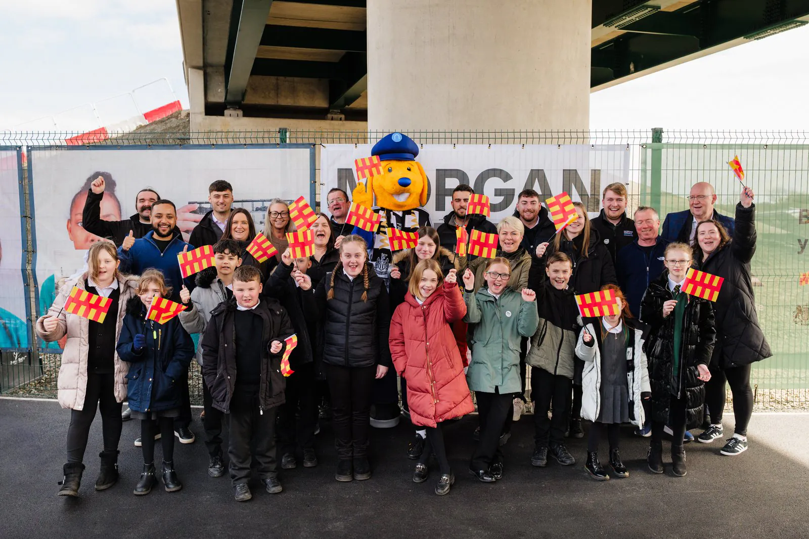 Group photo in front of a fence with banners, featuring people holding red and yellow flags and a bear mascot costume.