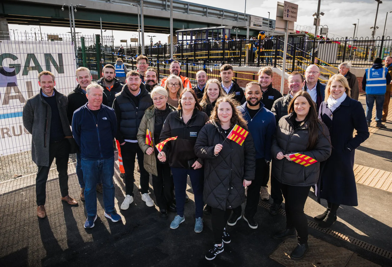 People gathered at a train station opening event with banners and signage in the background.