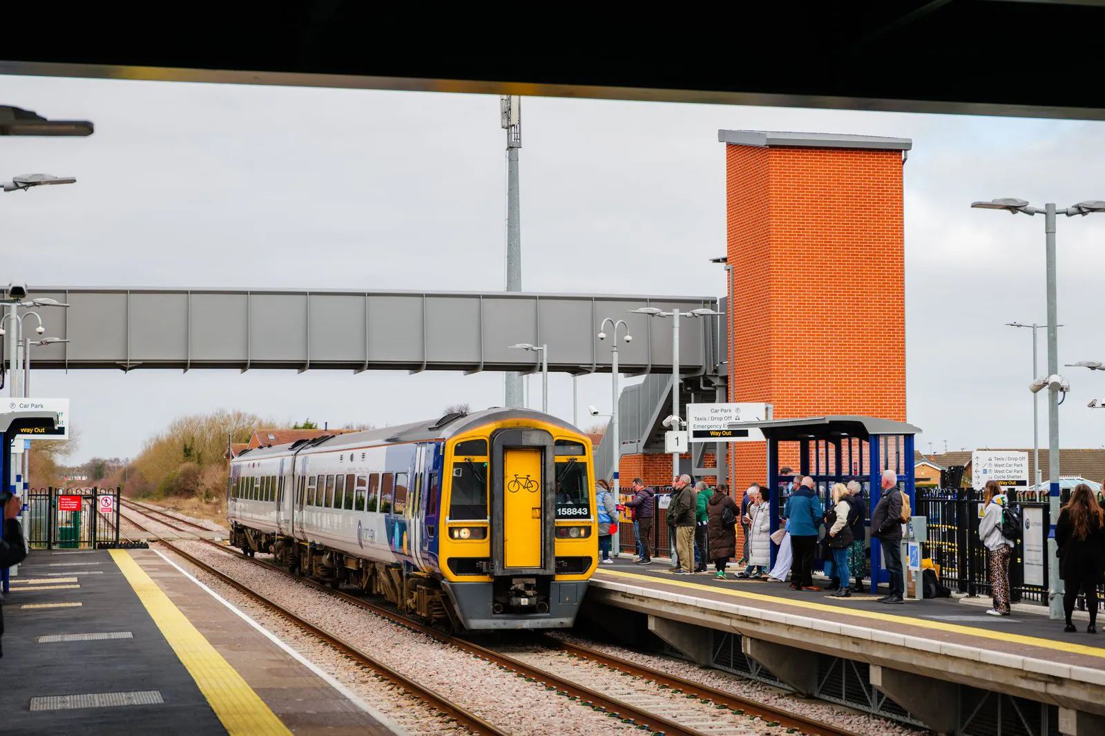 Train arrives or departs from a station platform with passengers waiting or boarding, and an overhead pedestrian bridge visible.