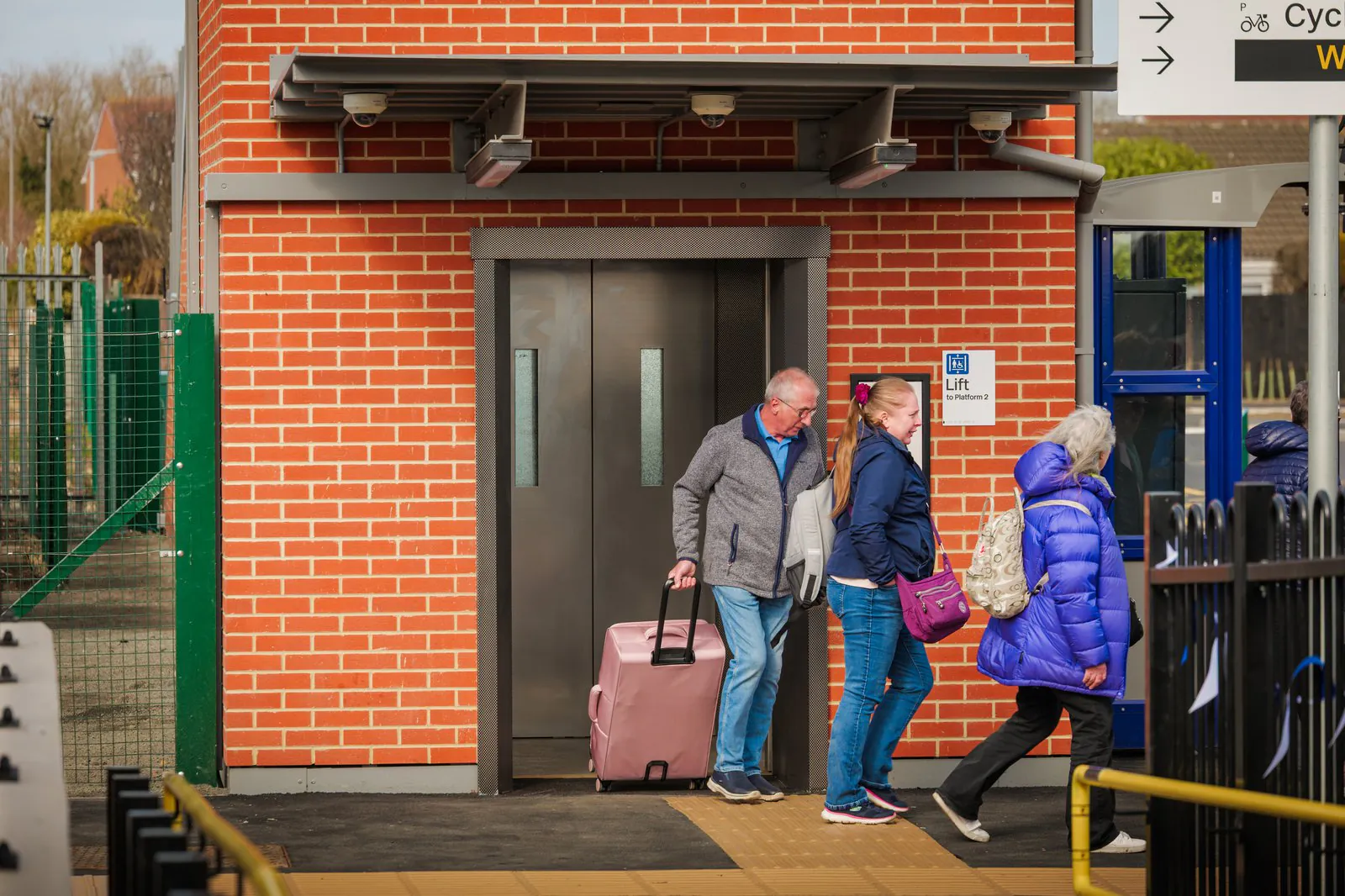 Three people with luggage stand outside an elevator at a train station, with accessibility signage and a brick building in the background.