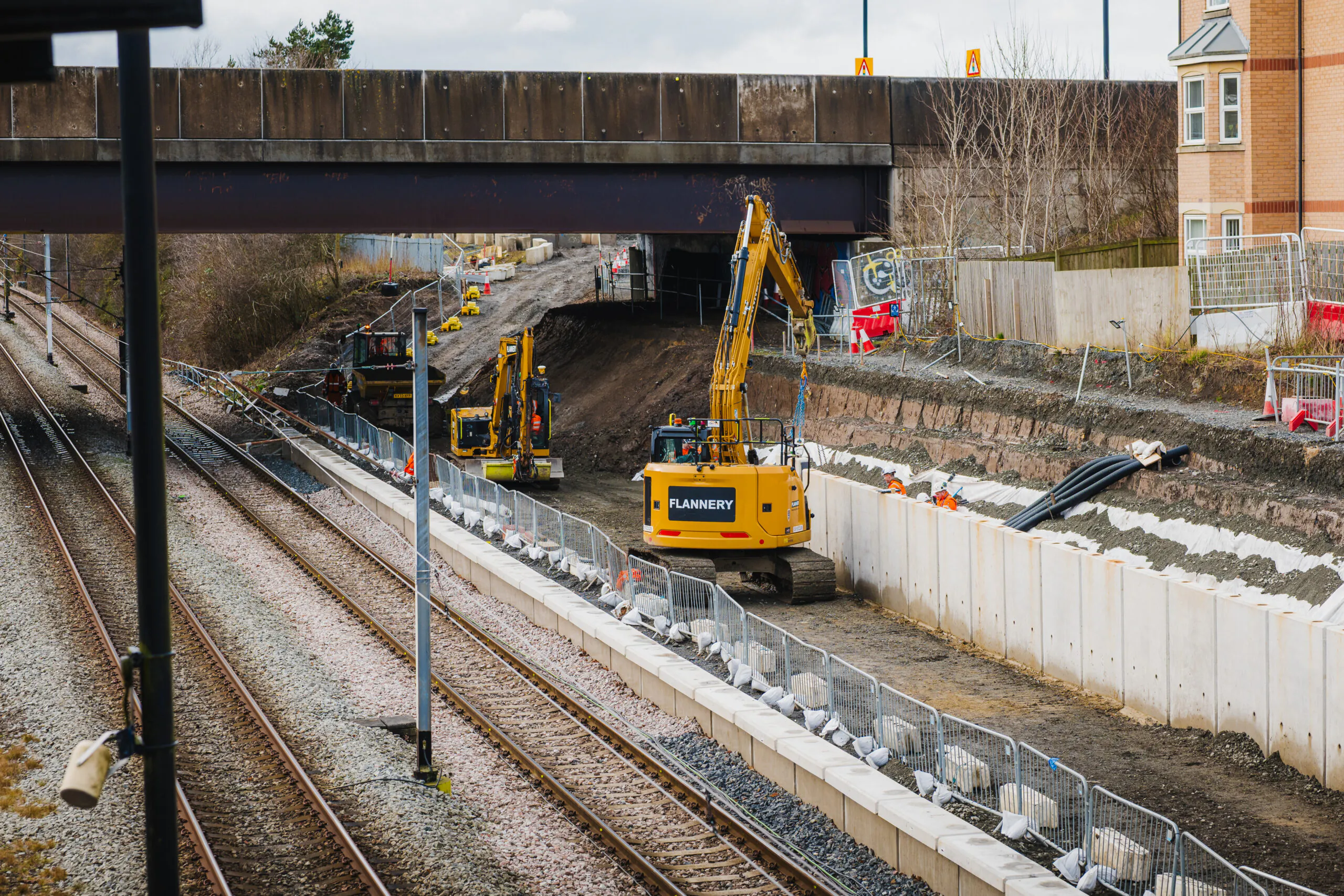 Excavator operating beside railway tracks during maintenance work, with workers in safety gear nearby.