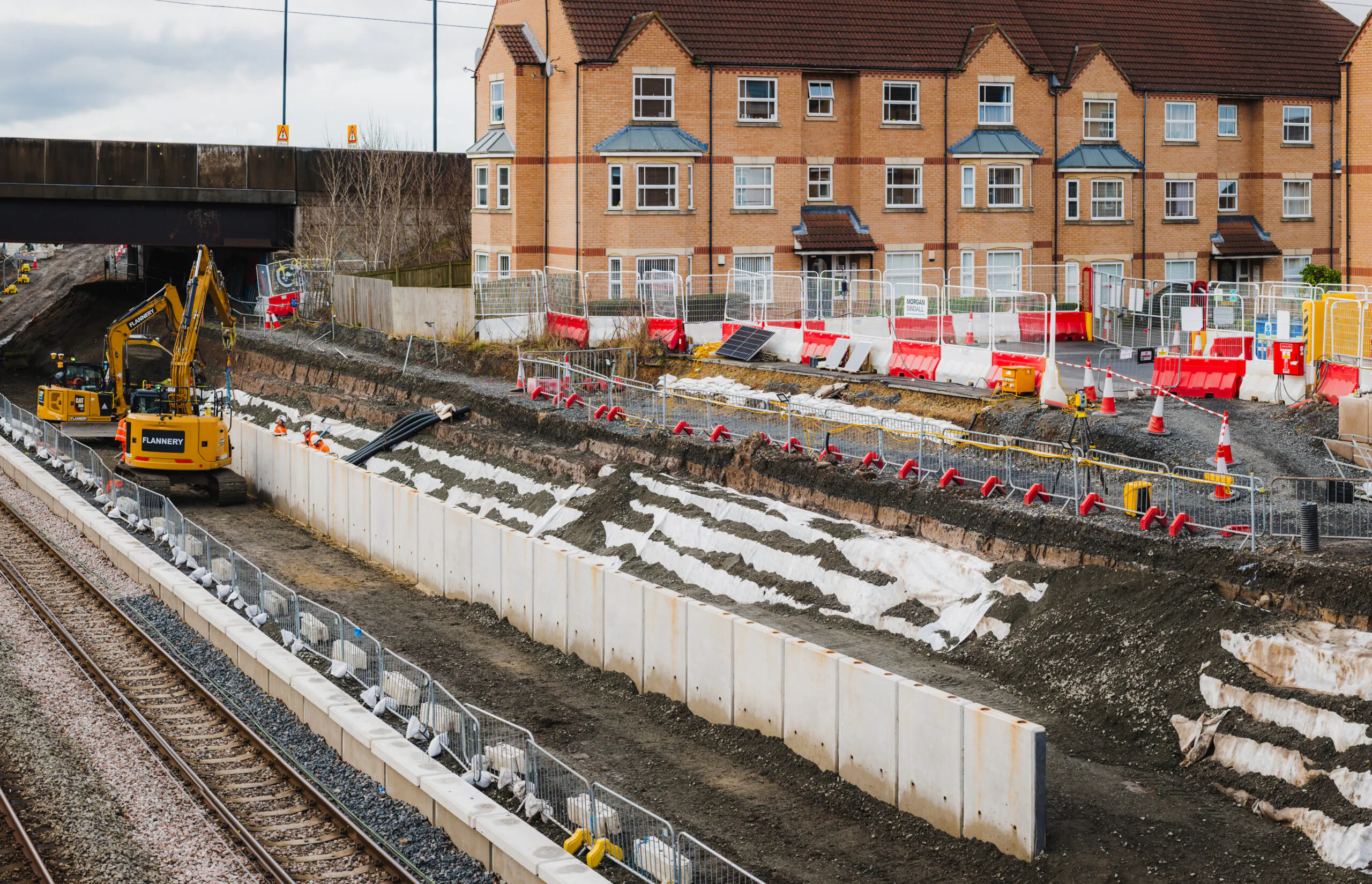 Excavator and workers performing maintenance beside railway tracks with safety barriers in place.