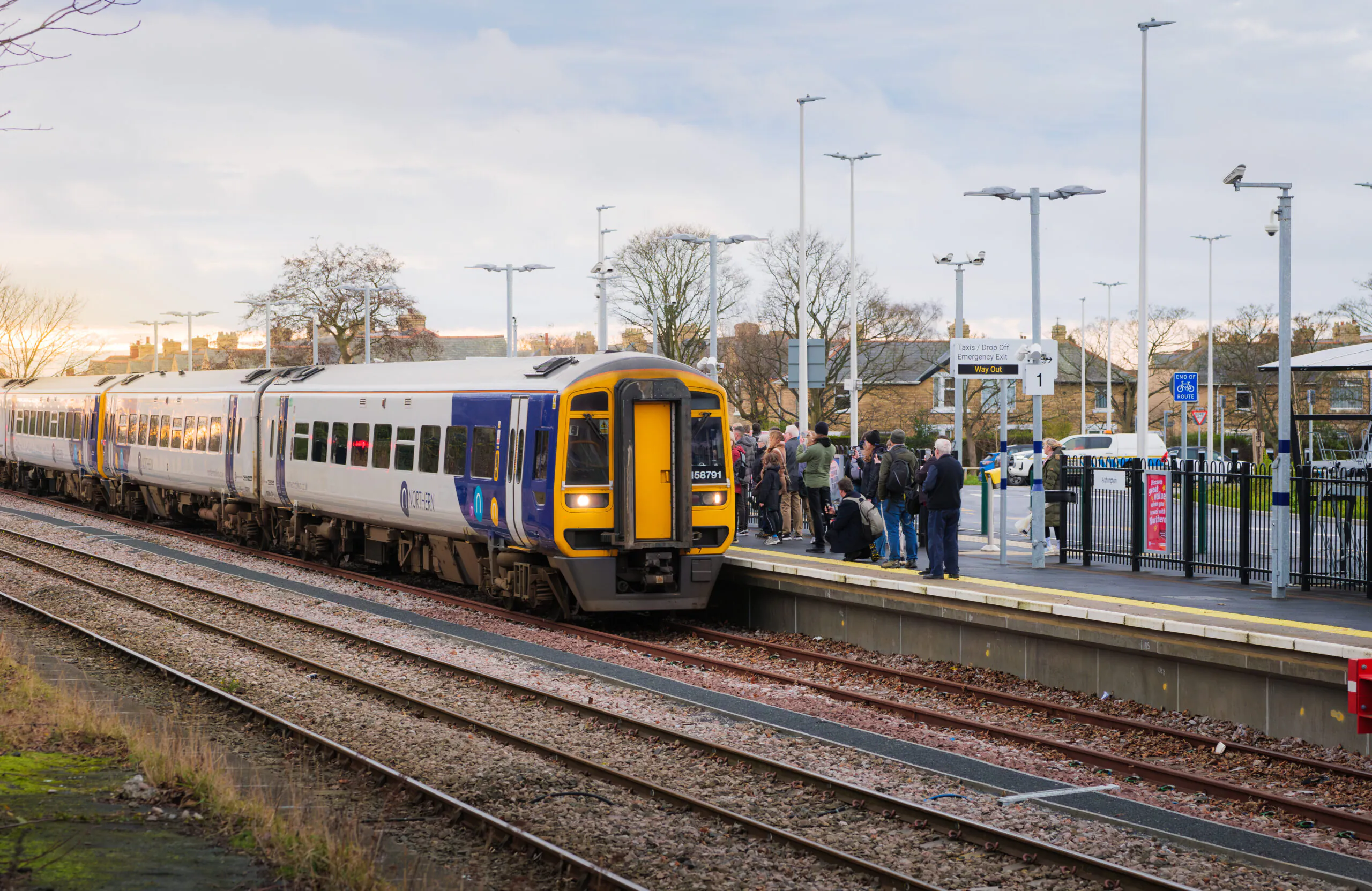 Train at a station platform with several people waiting, including passengers and workers in high-visibility clothing.