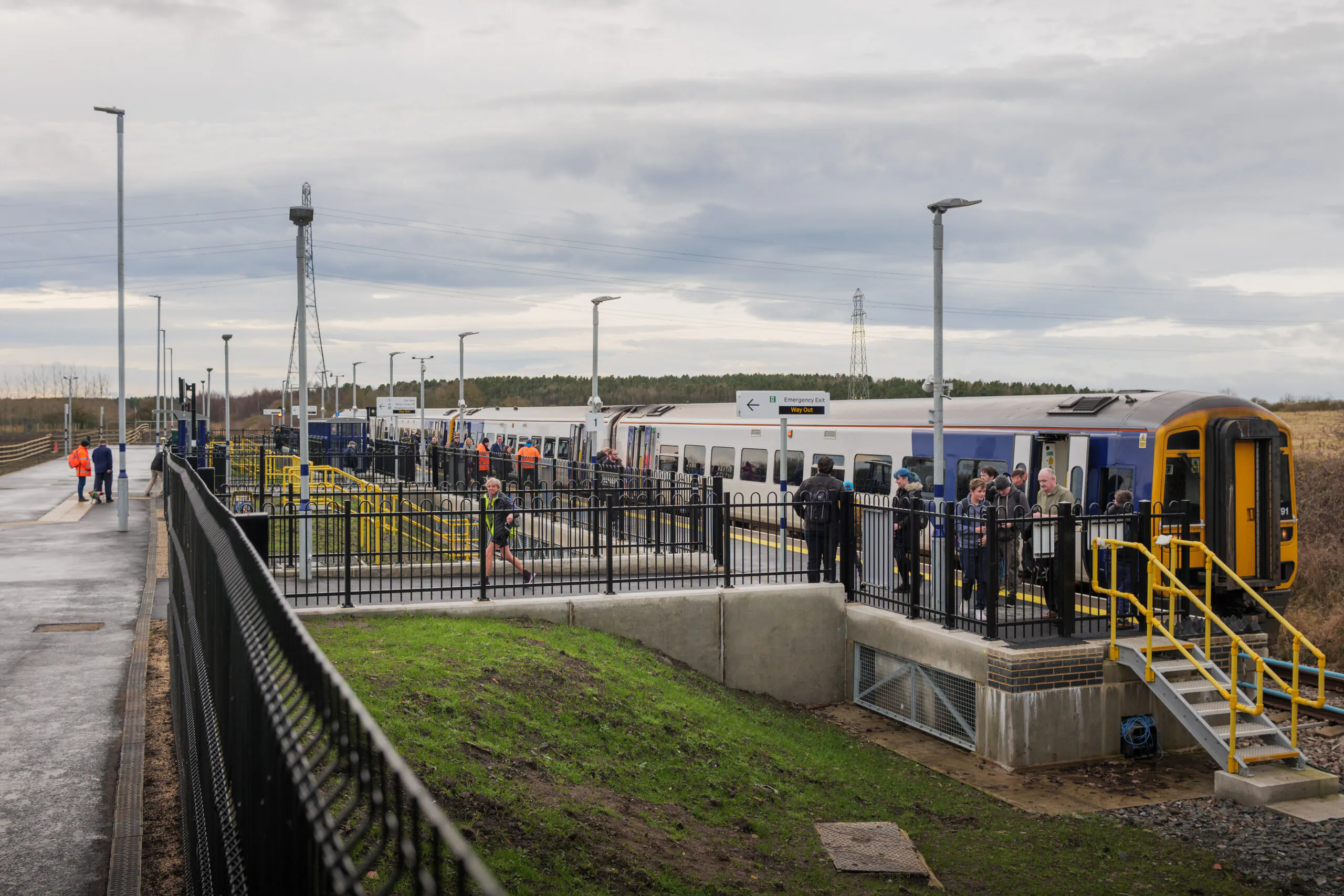 Train station platform with passengers boarding and walking along the platform under cloudy skies.