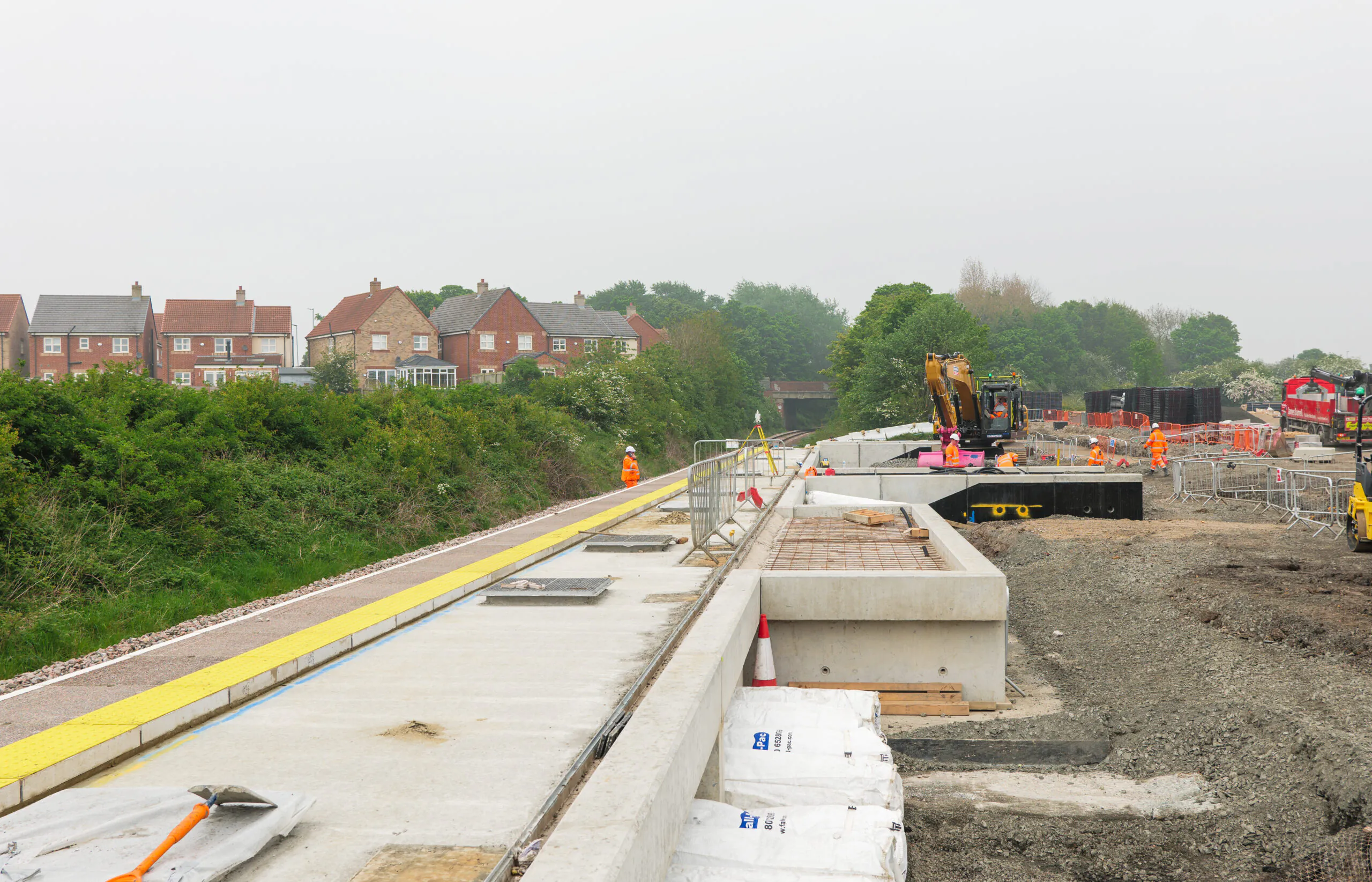 Construction site near residential houses with equipment and workers along an unfinished pathway or platform.