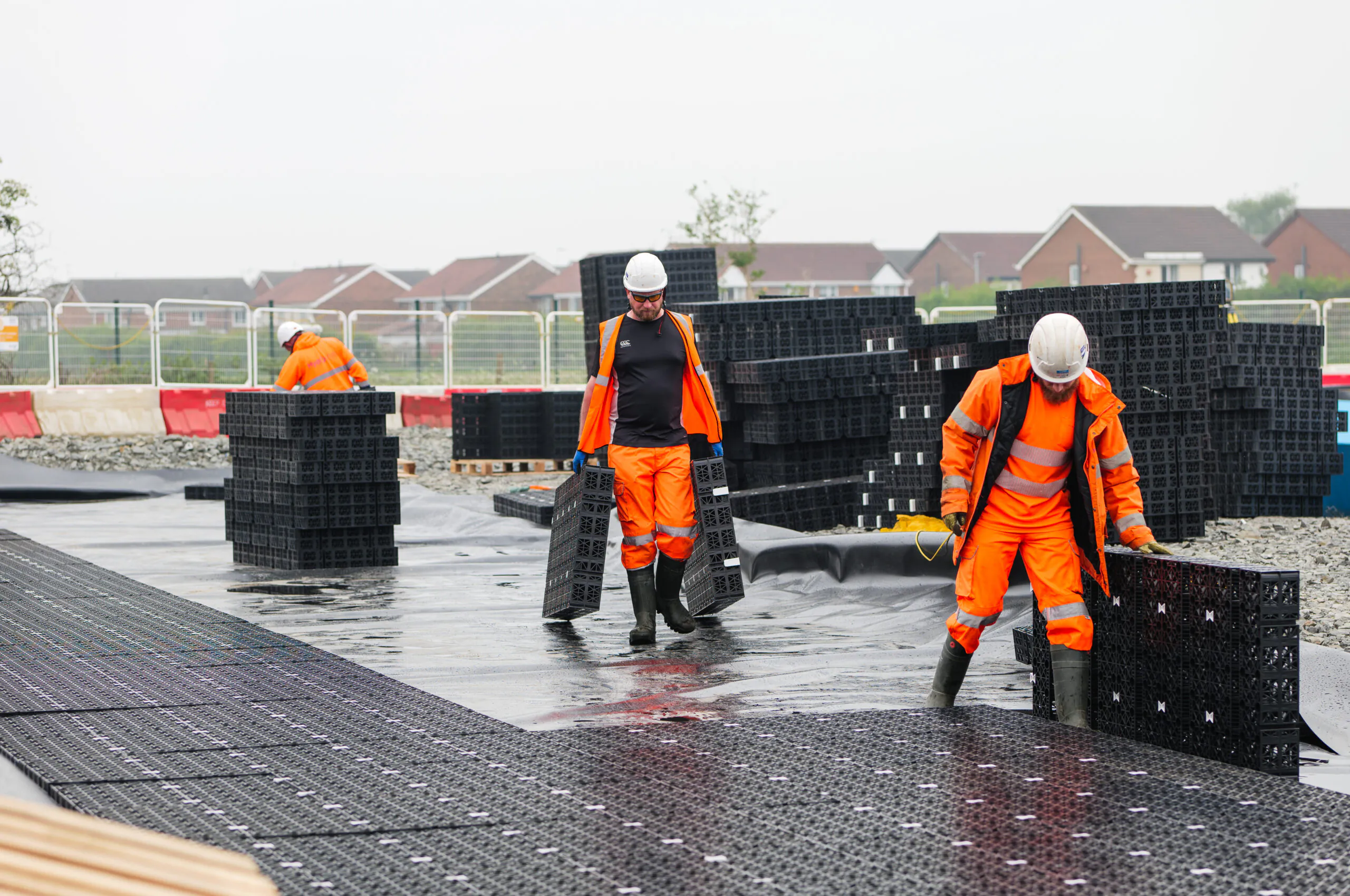 Workers in orange high-visibility clothing handling large black grid-like materials at a construction site near houses.