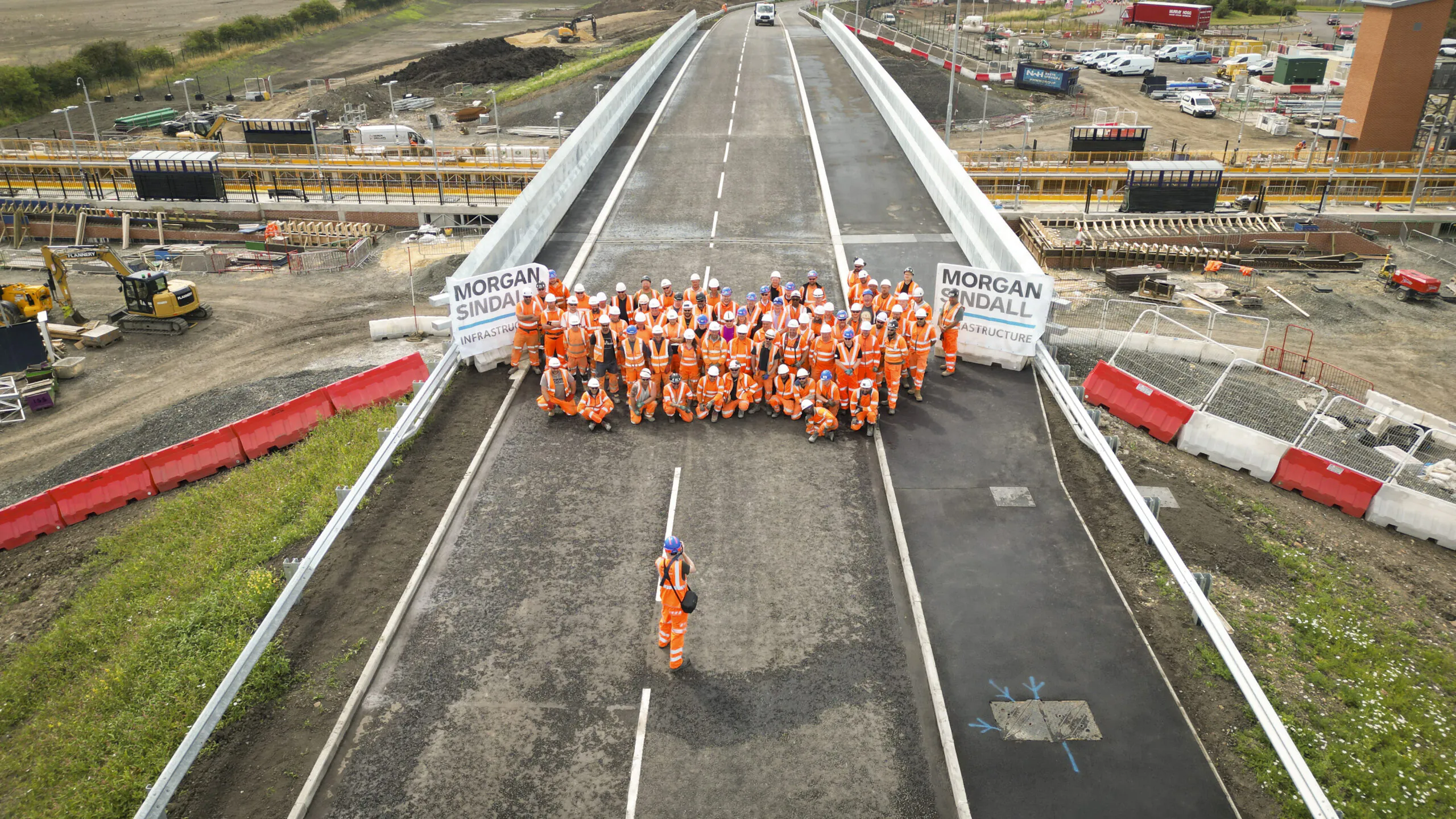 Large group of construction workers in orange safety gear stand on a newly built bridge with “Morgan Sindall” banners; construction machinery and materials are visible in the background.