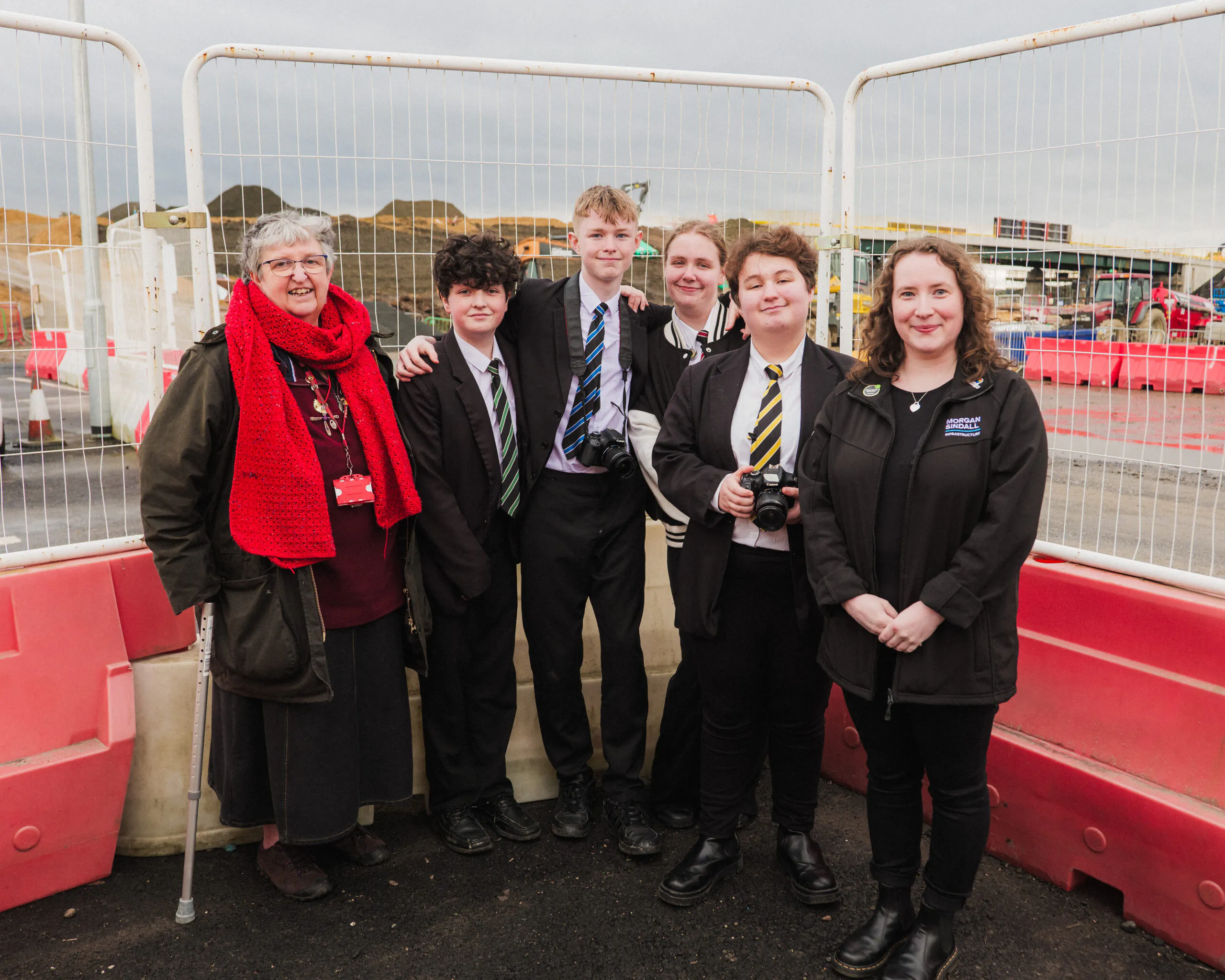 Group photo of six individuals standing behind temporary fencing at an active construction site.