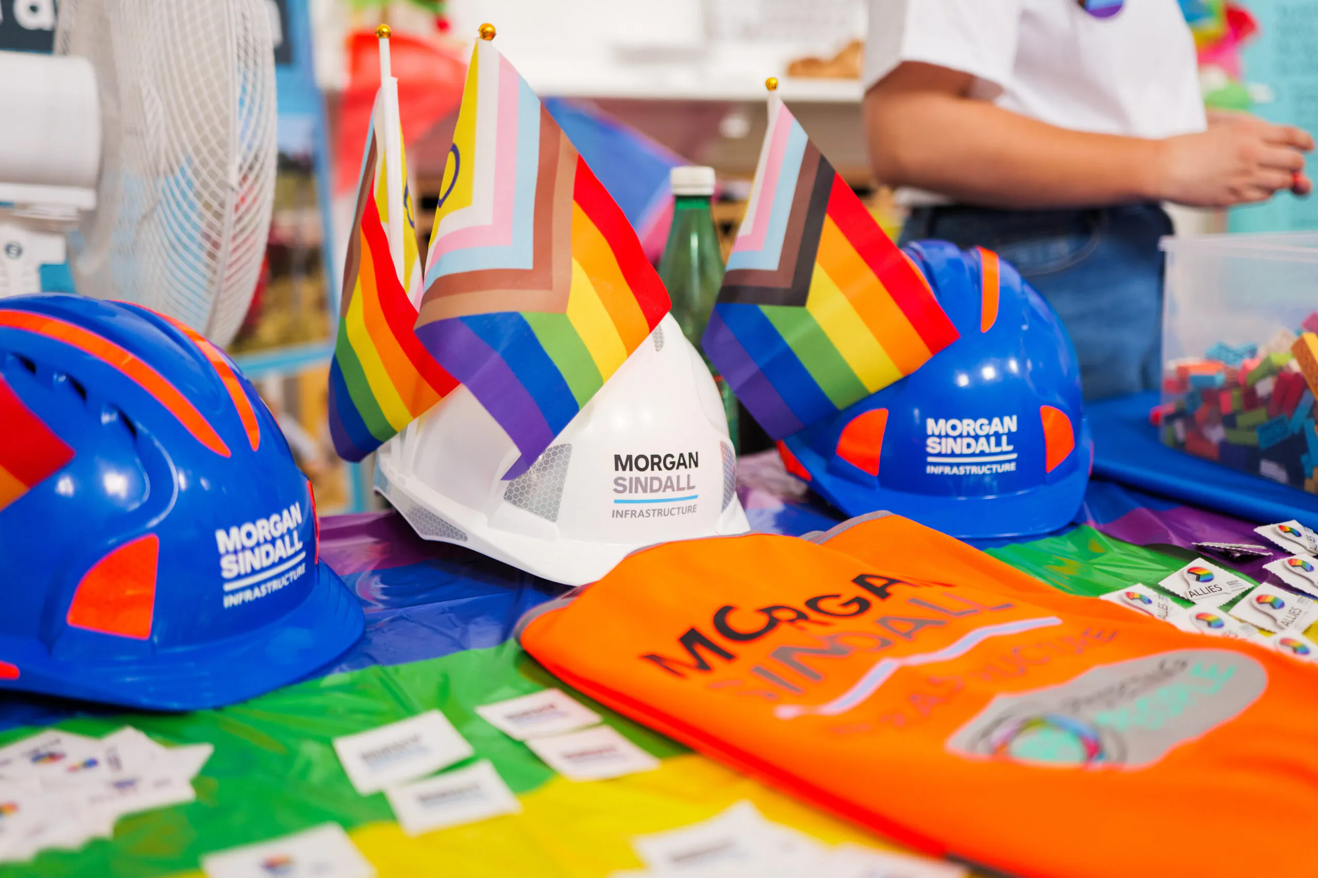 Display table with Morgan Sindall branded hard hats in blue and white, rainbow flags, and promotional items like stickers on a rainbow-themed tablecloth.