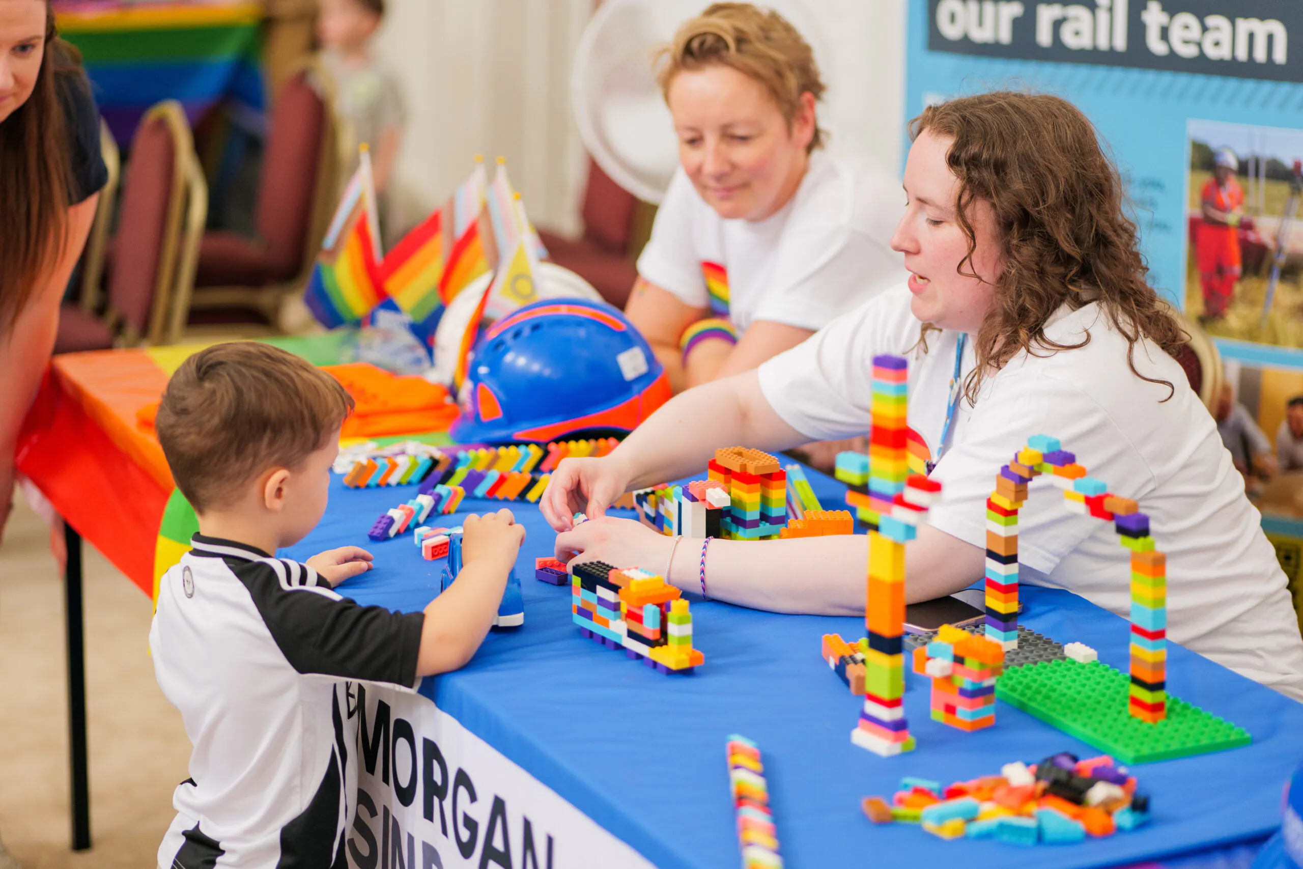 Child interacting with adults at an event booth featuring colorful LEGO structures and Morgan Sindall branding with rainbow flags promoting diversity.