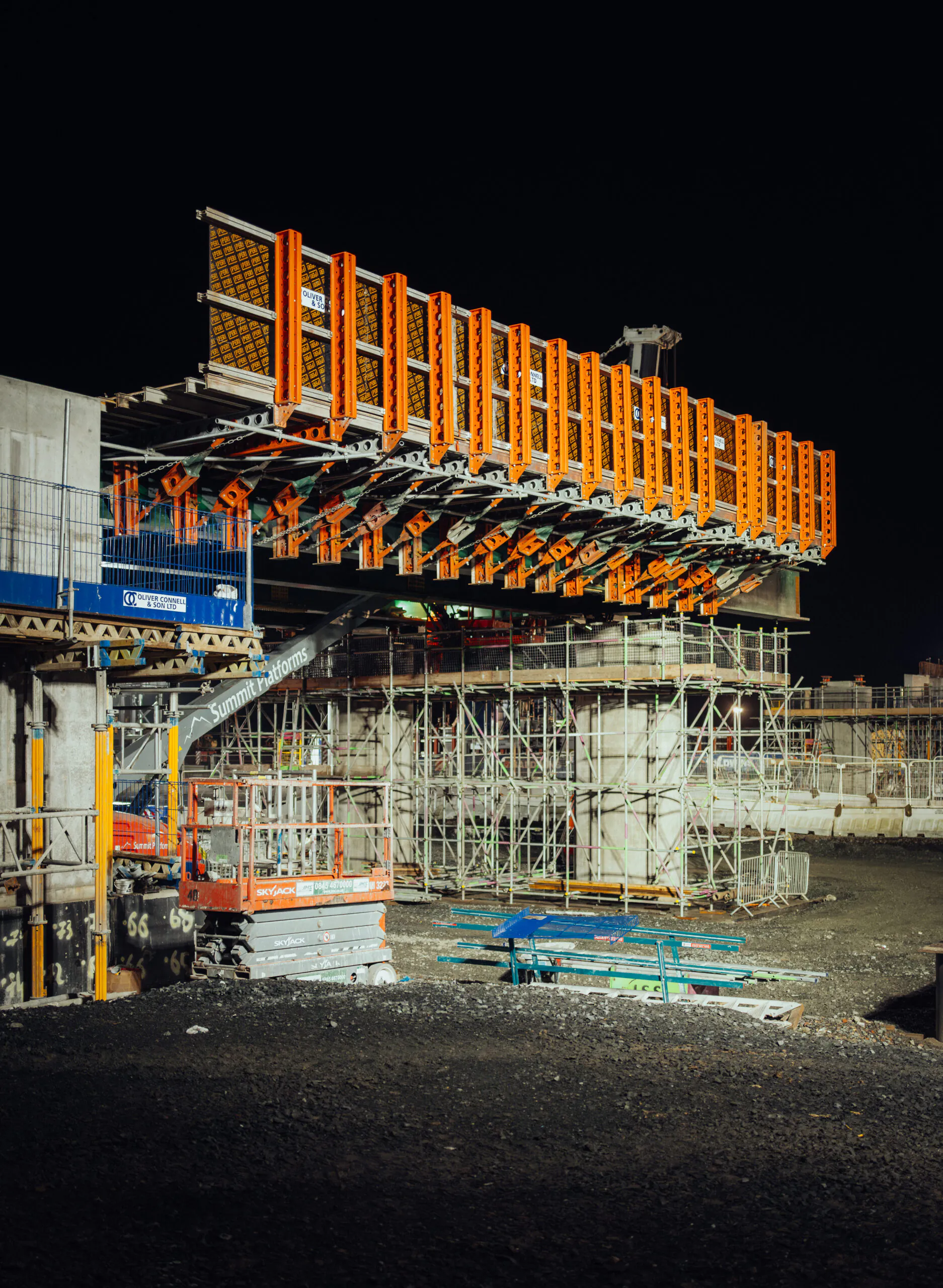 Nighttime construction site with a partially built bridge supported by scaffolding and bright orange components.