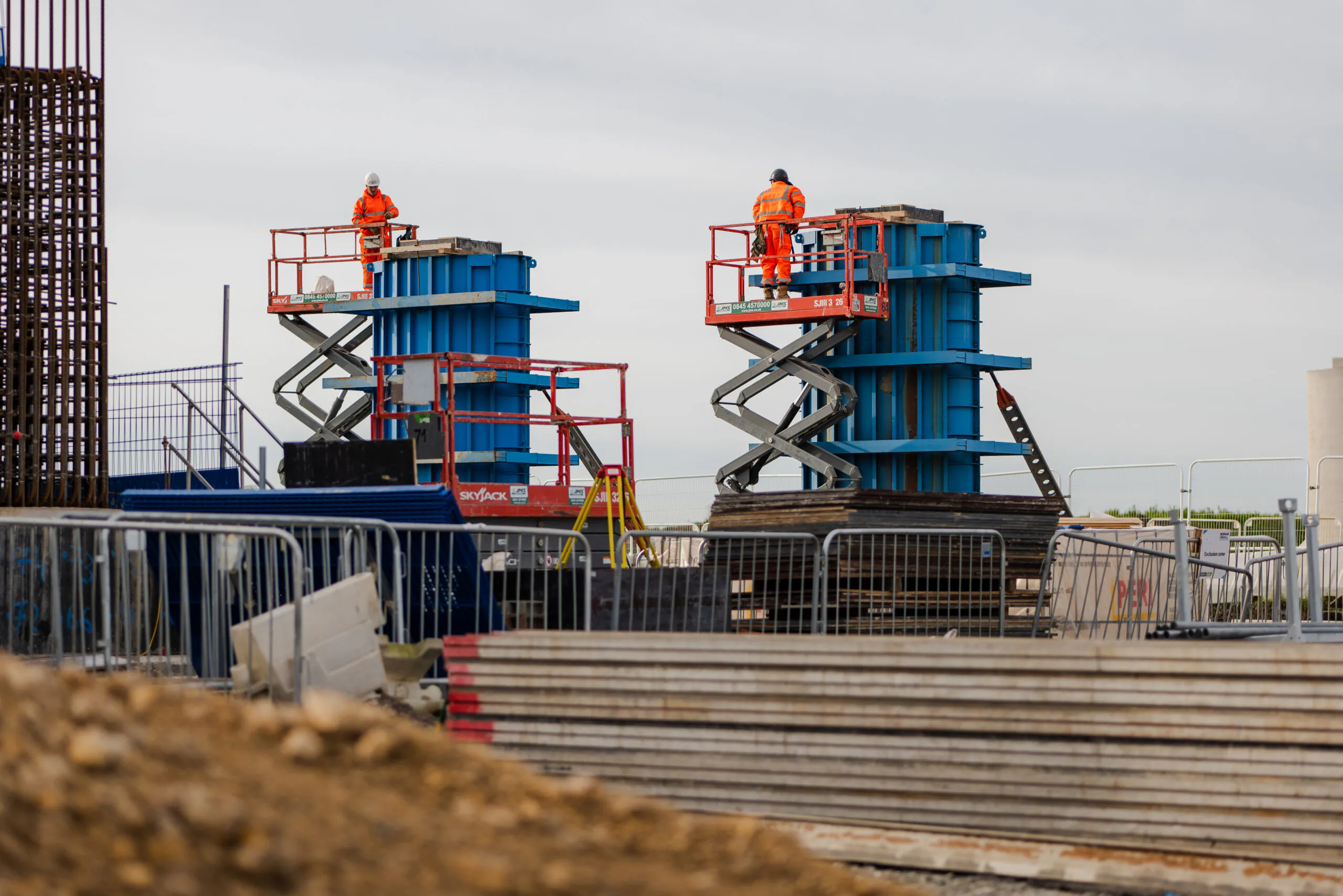 Two workers operating scissor lifts next to blue structural columns at an outdoor construction site during daylight.