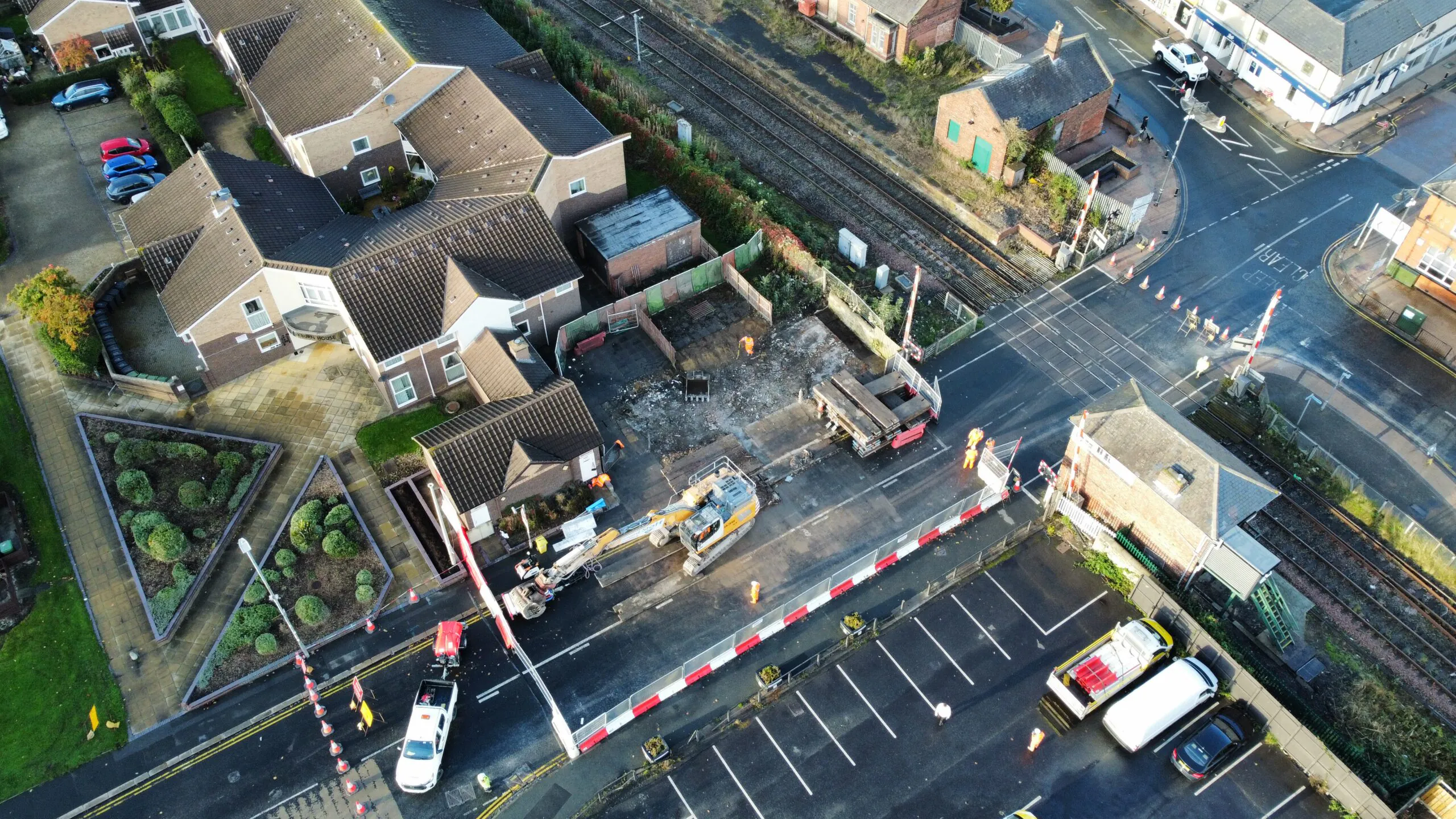 Aerial view of a construction site near railway tracks and residential houses, with vehicles, workers, and safety barriers visible.