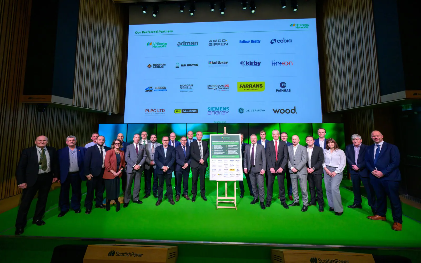 Group of formally dressed individuals stand on a green stage with a large screen showing company logos, at an SP Energy Networks event.