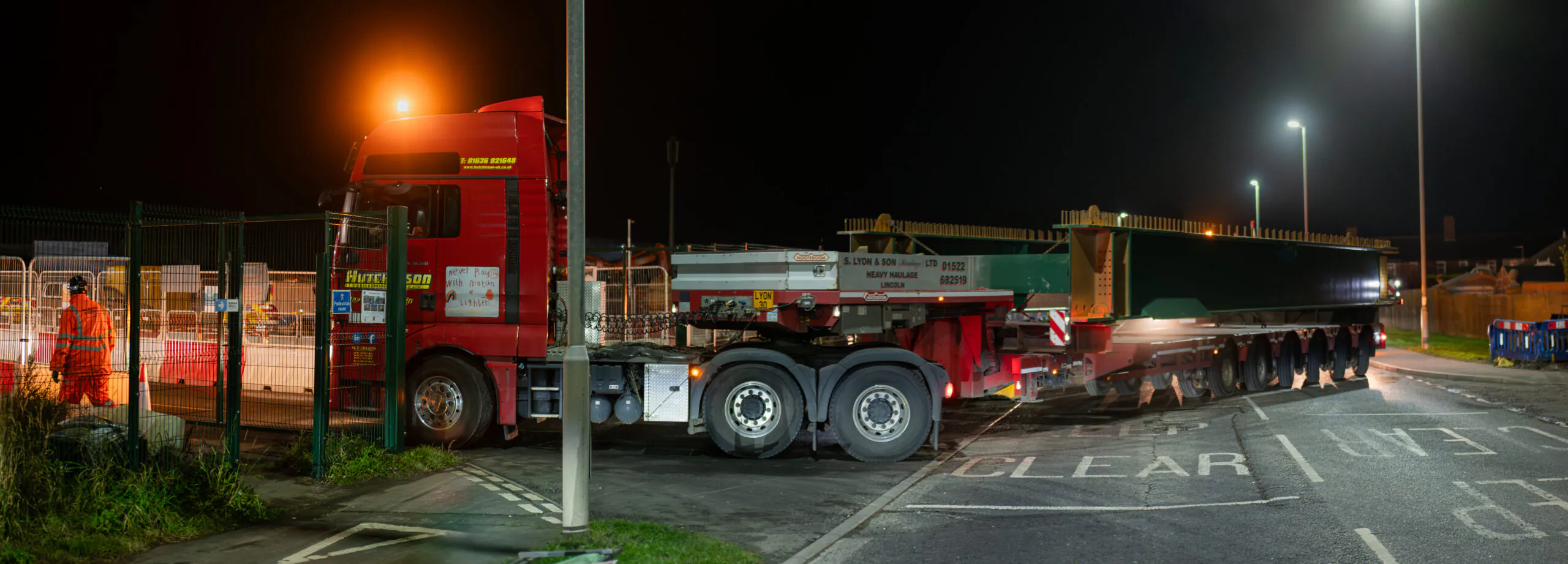Large red truck with a flatbed trailer carrying a green steel beam at night near a fenced construction area.