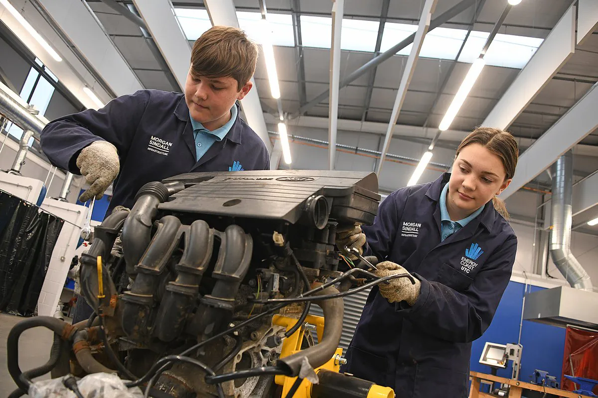 Two individuals in blue work uniforms and gloves work on an engine inside an industrial facility.