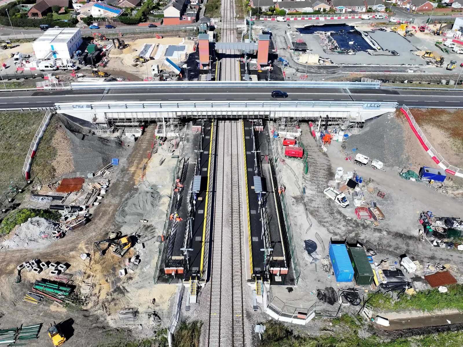 Aerial view of a construction site with roads, buildings, and surrounding green fields under clear skies.