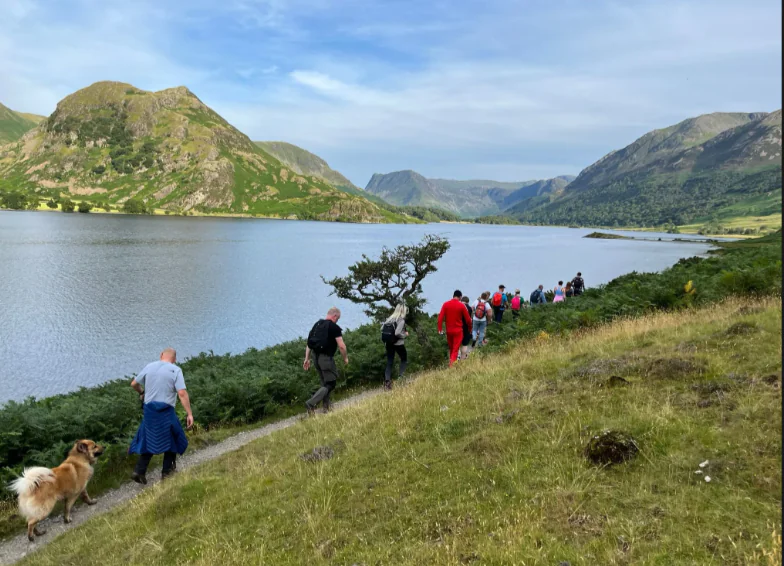 Group of hikers walk beside a lake surrounded by hills and mountains under clear skies; one hiker is accompanied by a dog.