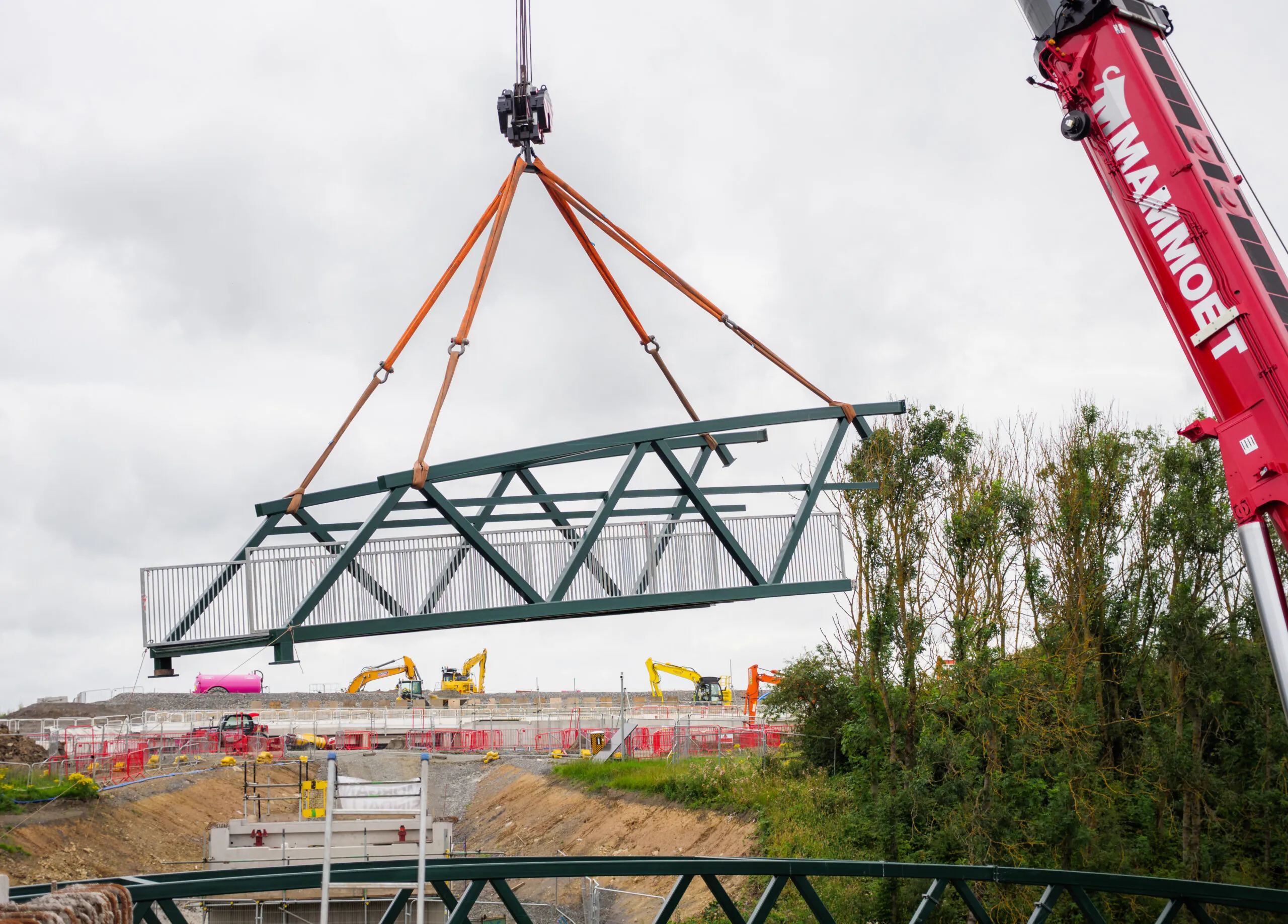 Large steel bridge section being lifted by cranes at a construction site with heavy machinery in the background.