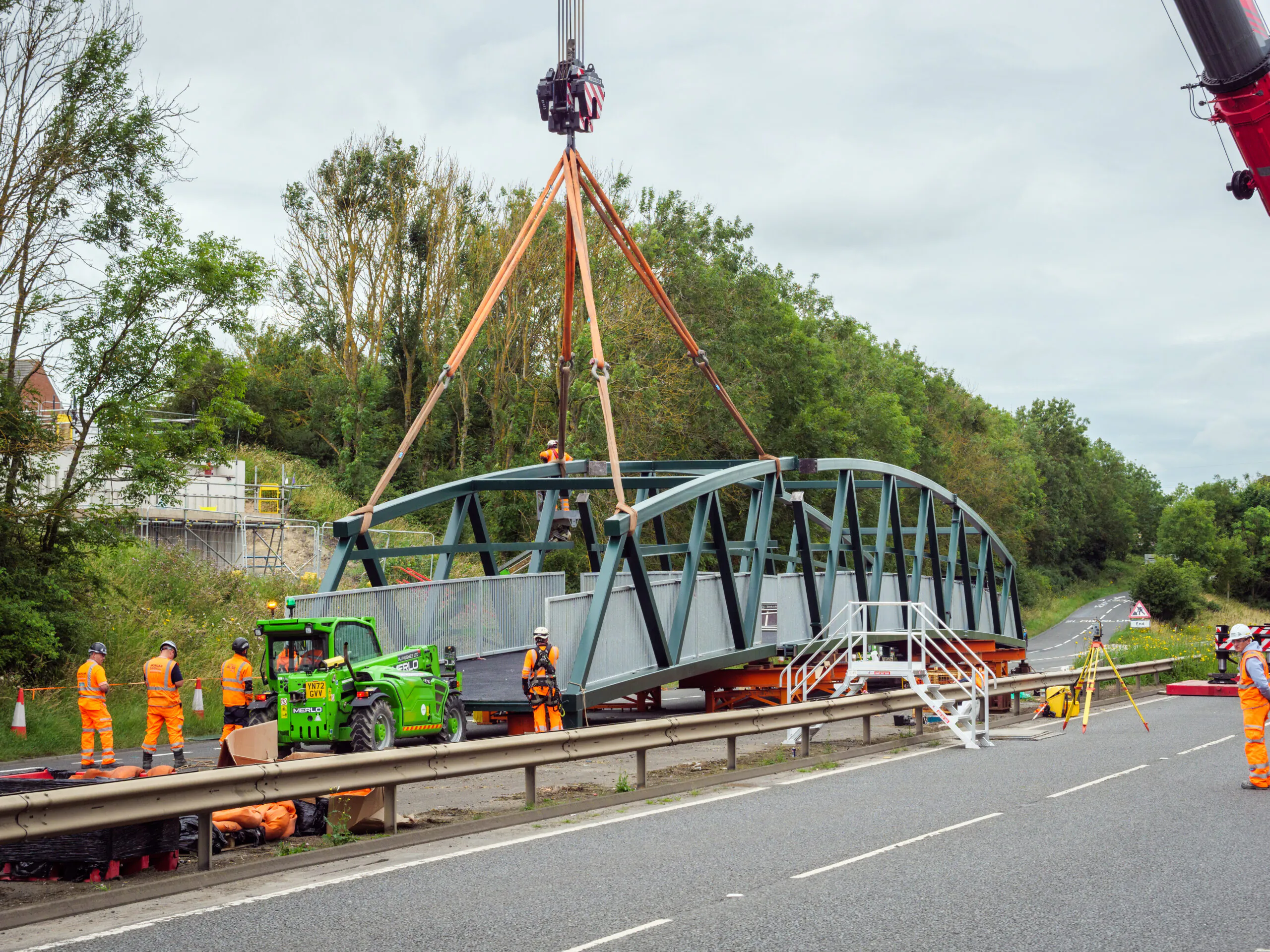 Steel bridge section being maneuvered by cranes at a construction site with workers and vehicles nearby.