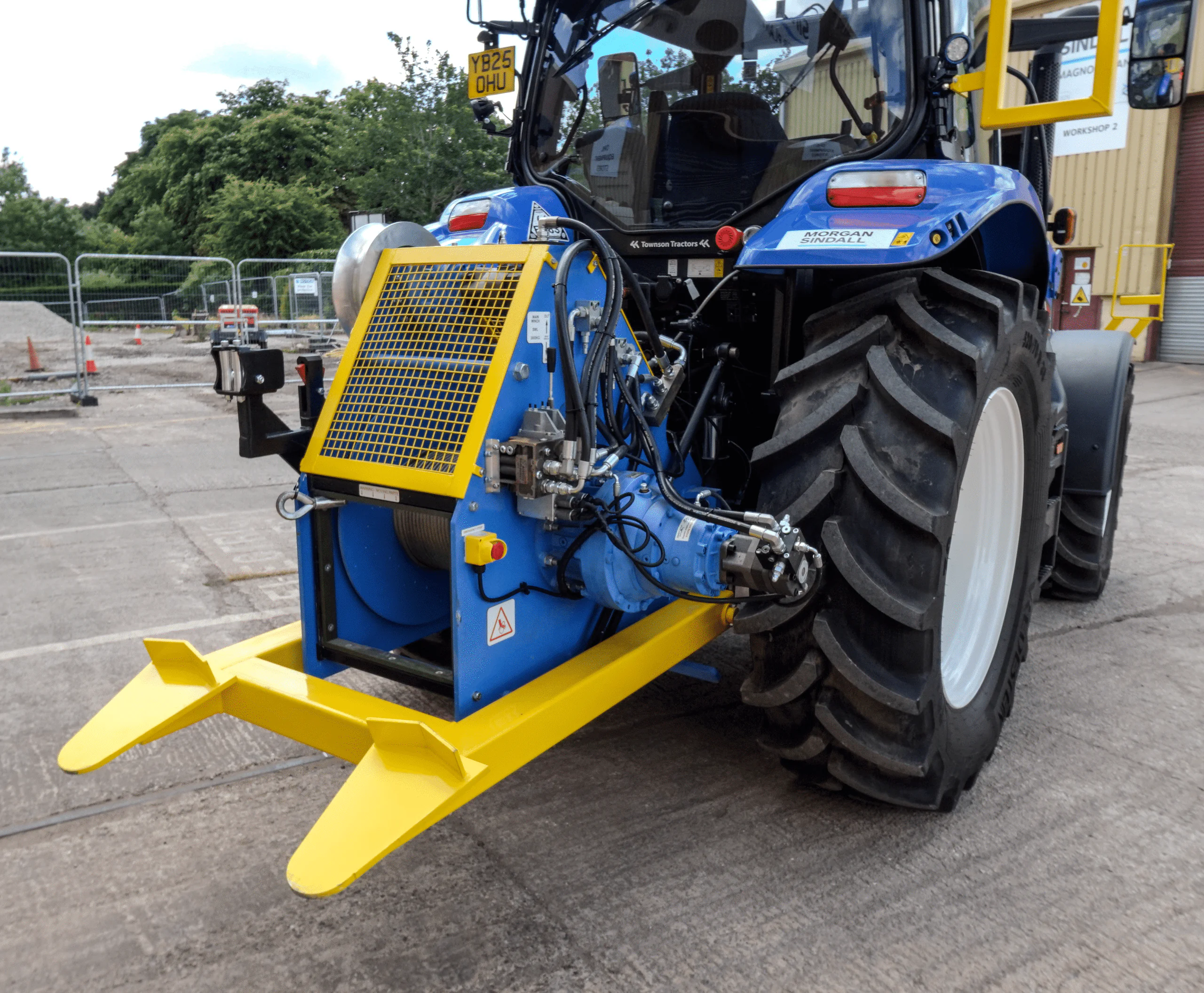 Close-up of heavy machinery mounted on the back of a blue tractor parked near fencing.