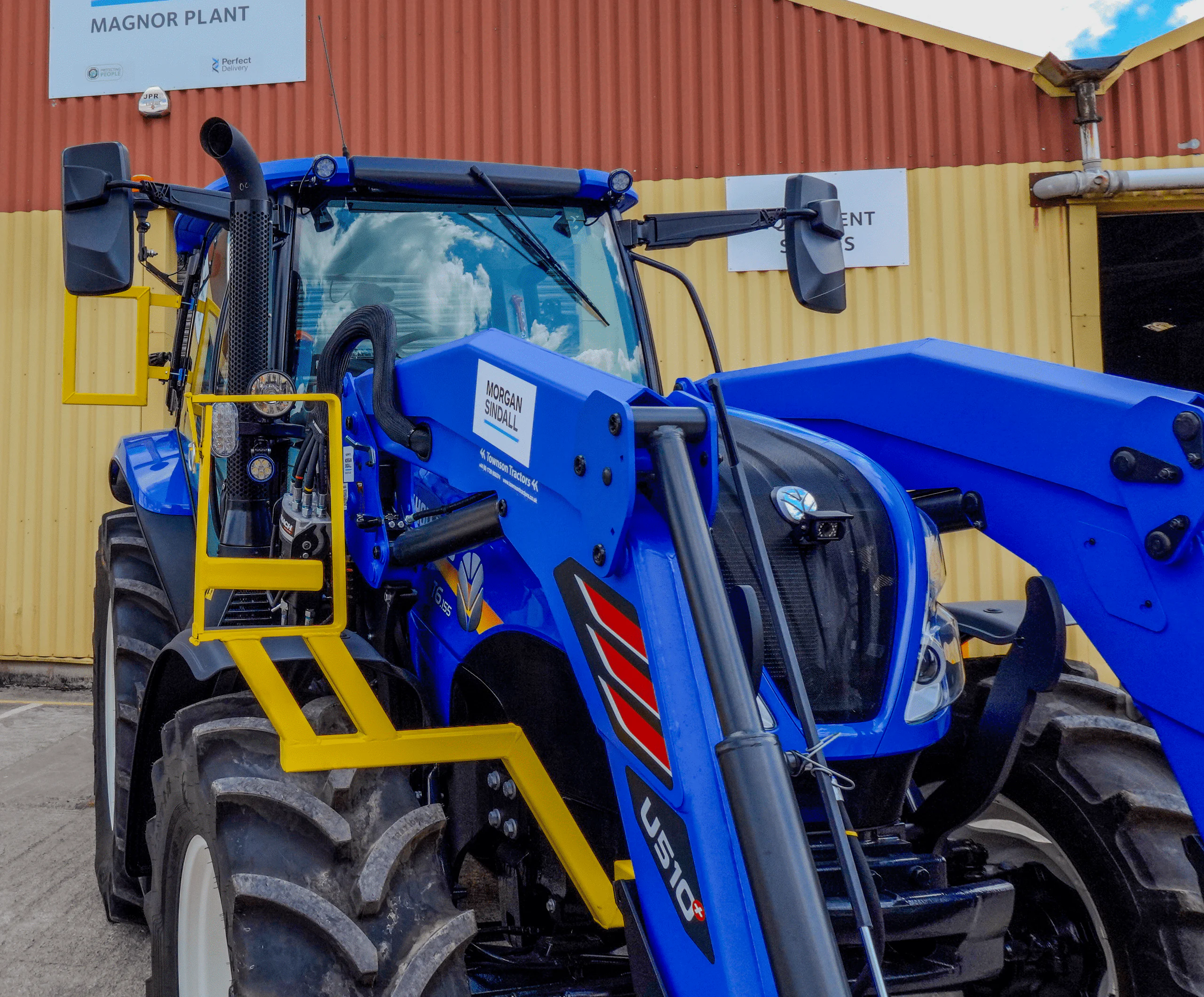 Blue tractor with specialized attachments parked outside a building labeled Magnor Plant Equipment Services.