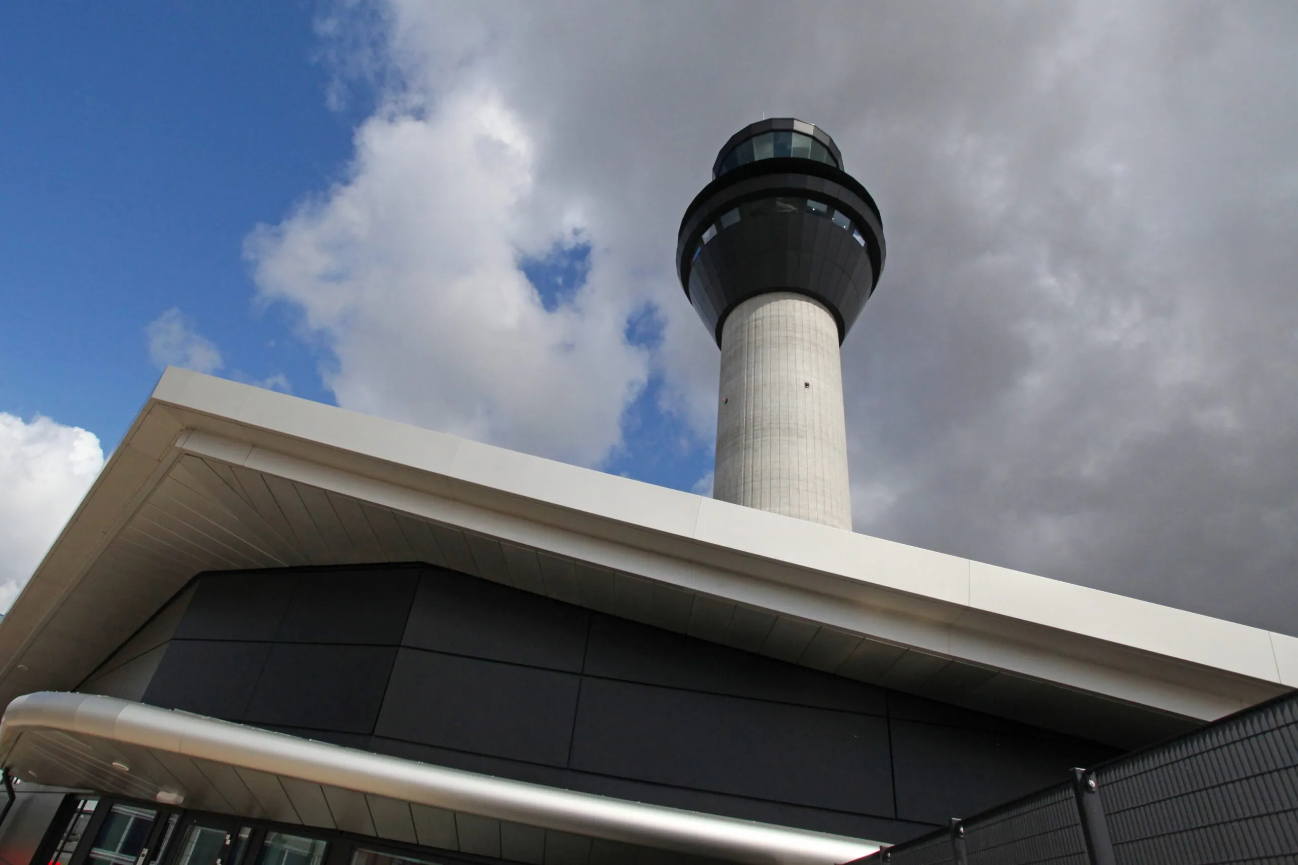 Ground-level view of an airport control tower with fire trucks parked nearby inside a fenced area under partly cloudy skies.