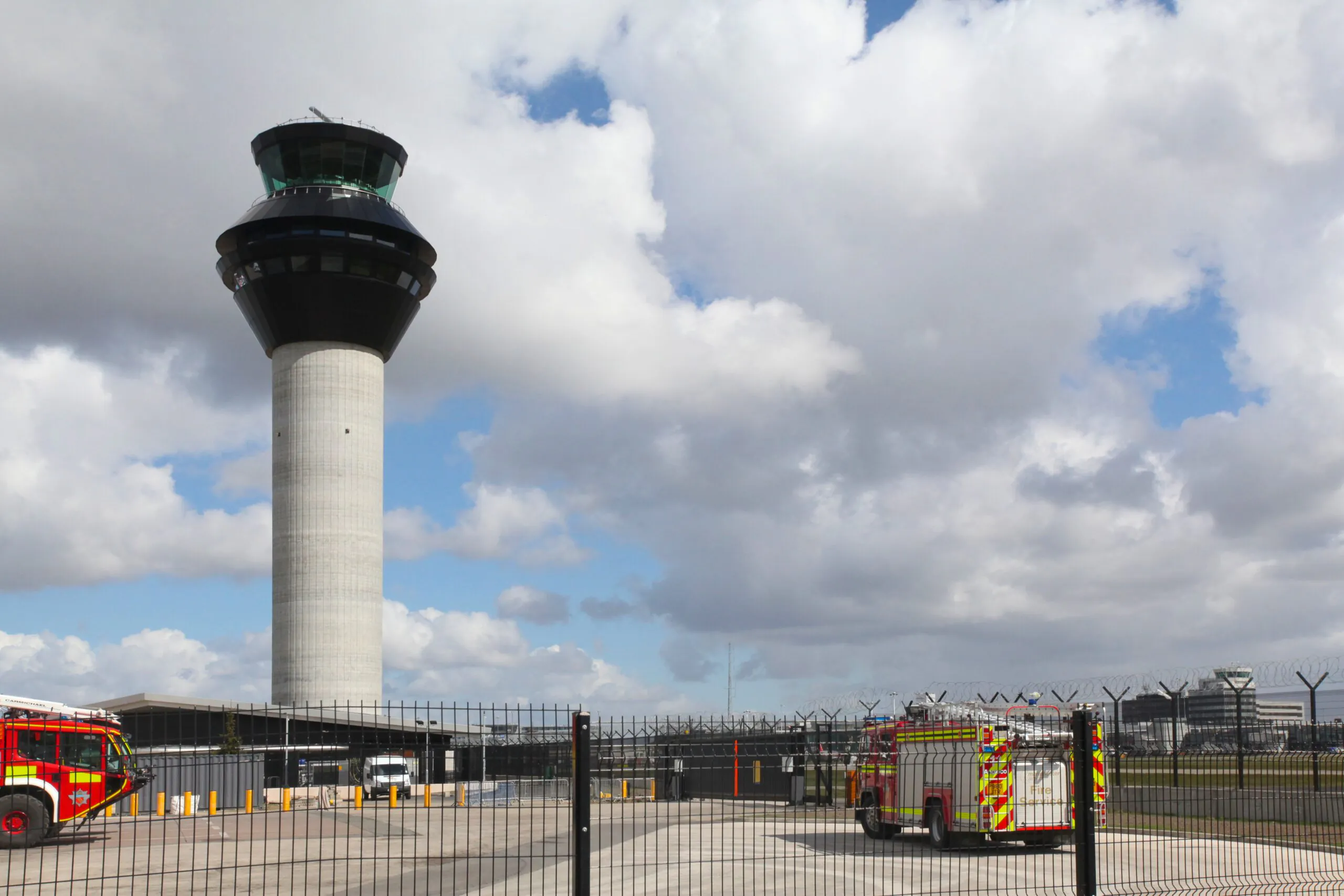 Ground-level view of an airport control tower with fire trucks parked nearby inside a fenced area under partly cloudy skies.