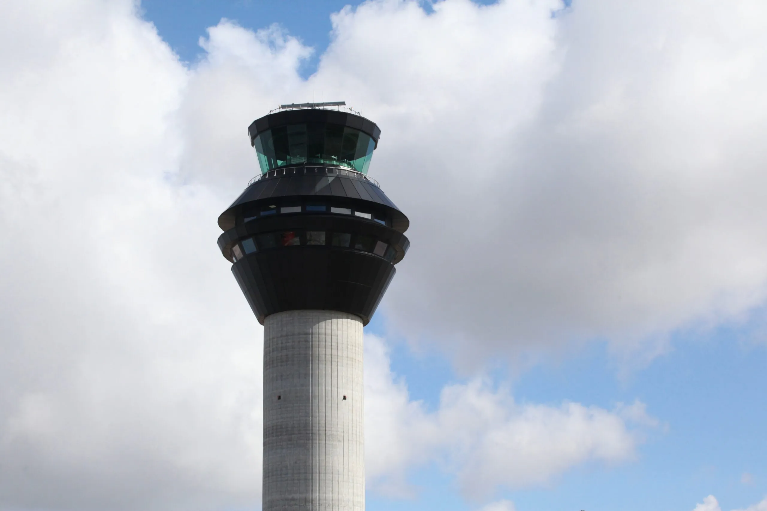 Ground-level view of an airport control tower with fire trucks parked nearby inside a fenced area under partly cloudy skies.