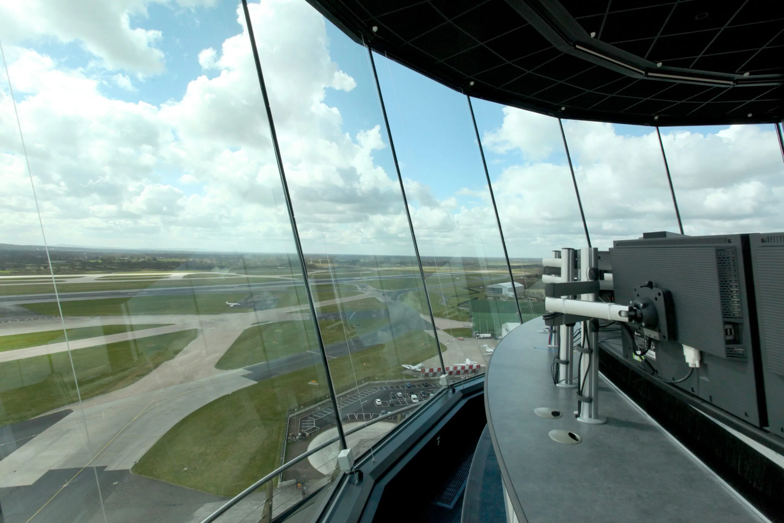 Interior of an airport control tower showing equipment setup and panoramic windows overlooking runways under partly cloudy skies.