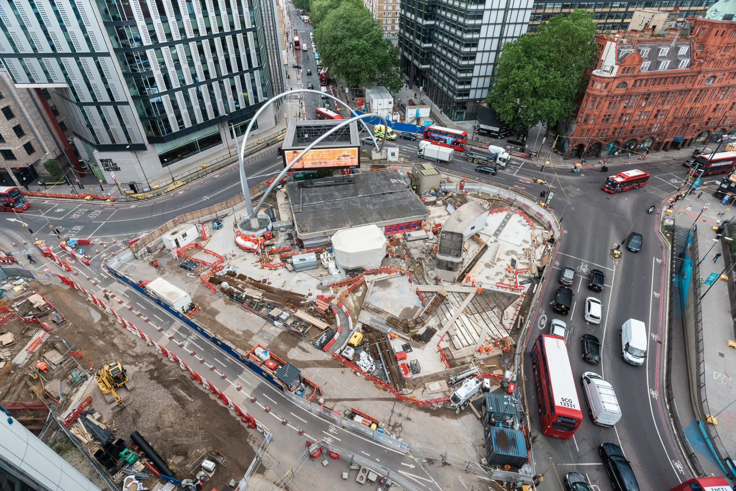 Aerial view of an urban construction site surrounded by busy roads with vehicles and buildings; red-and-white barriers mark the work zone.