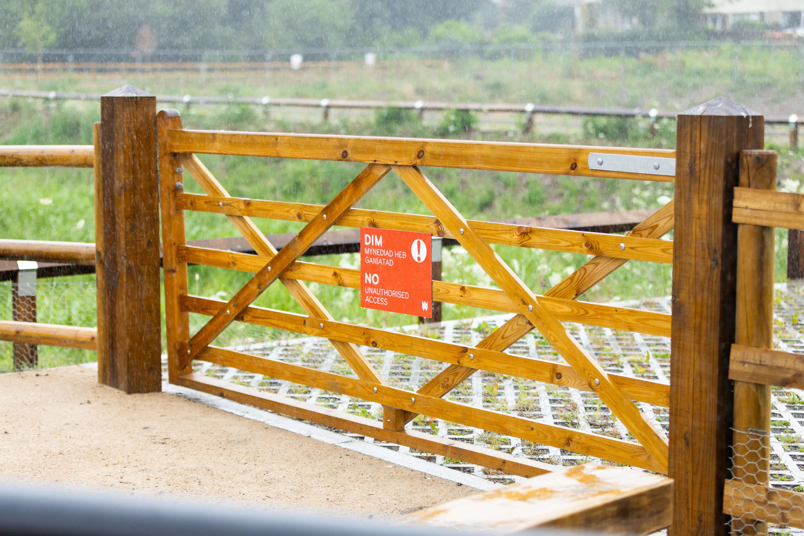 Wooden gate with diagonal crossbars and red sign reading "DIM MYNEDIAD HEB AWDLAD NO UNAUTHORISED ACCESS."