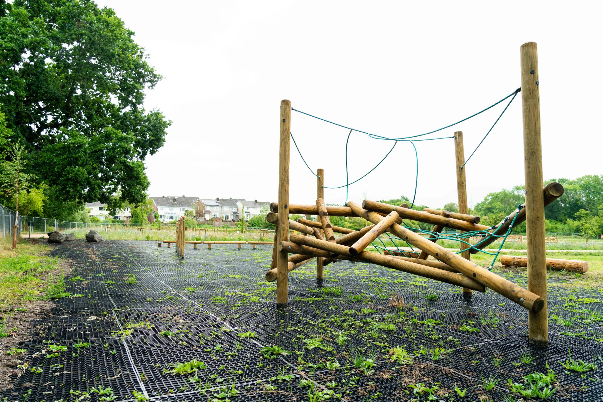 Wooden climbing structure with ropes on a black grid surface in an open area near houses and trees.