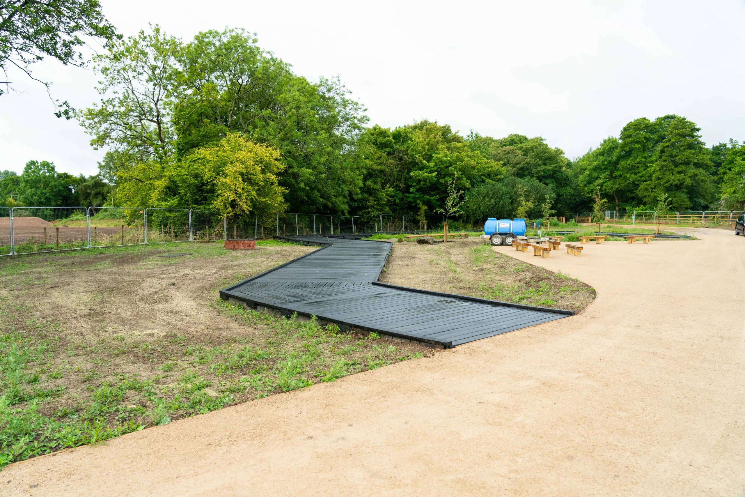 Landscaped area with newly planted trees and greenery along a pathway made of black plastic panels, with barrels placed nearby.