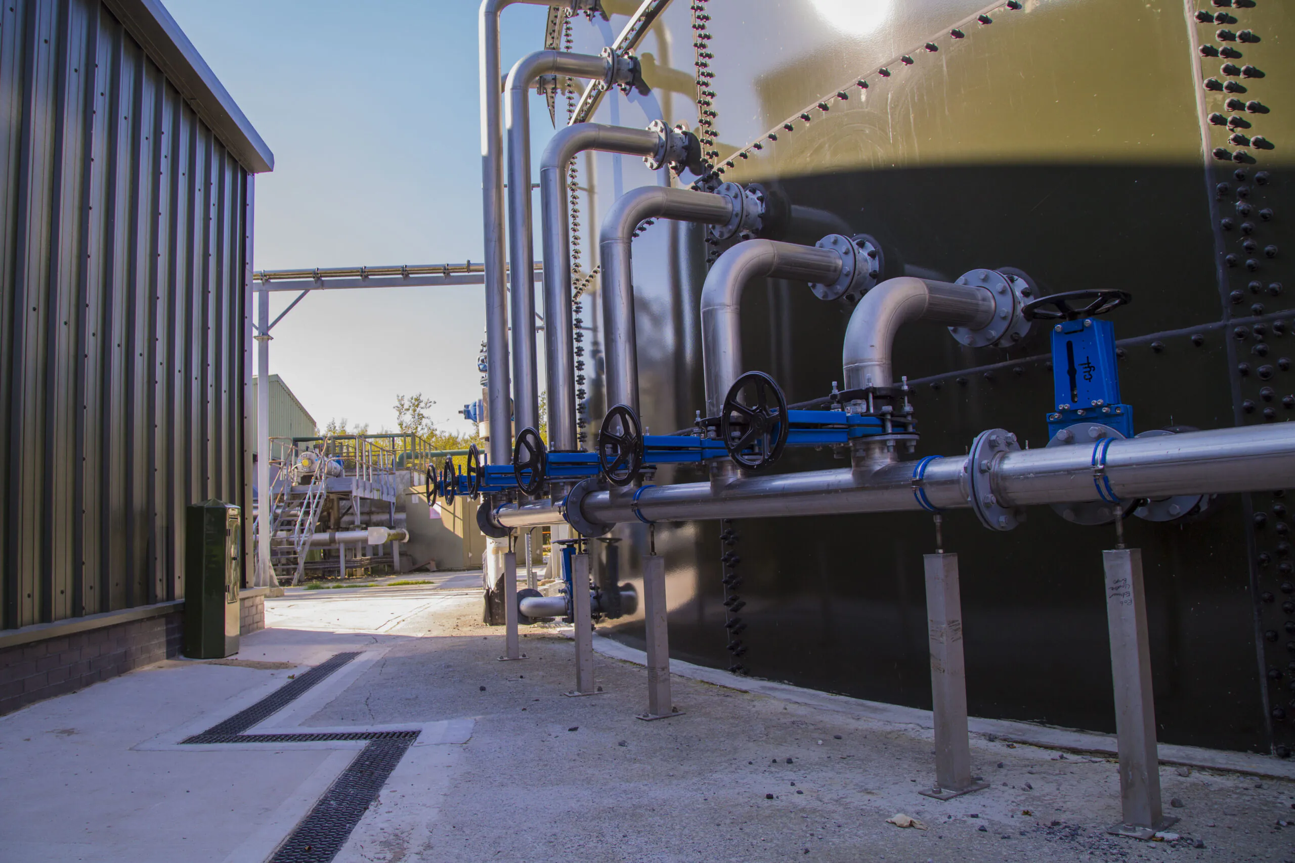 Industrial pipes and blue valves connected to large metallic tanks at an outdoor facility.